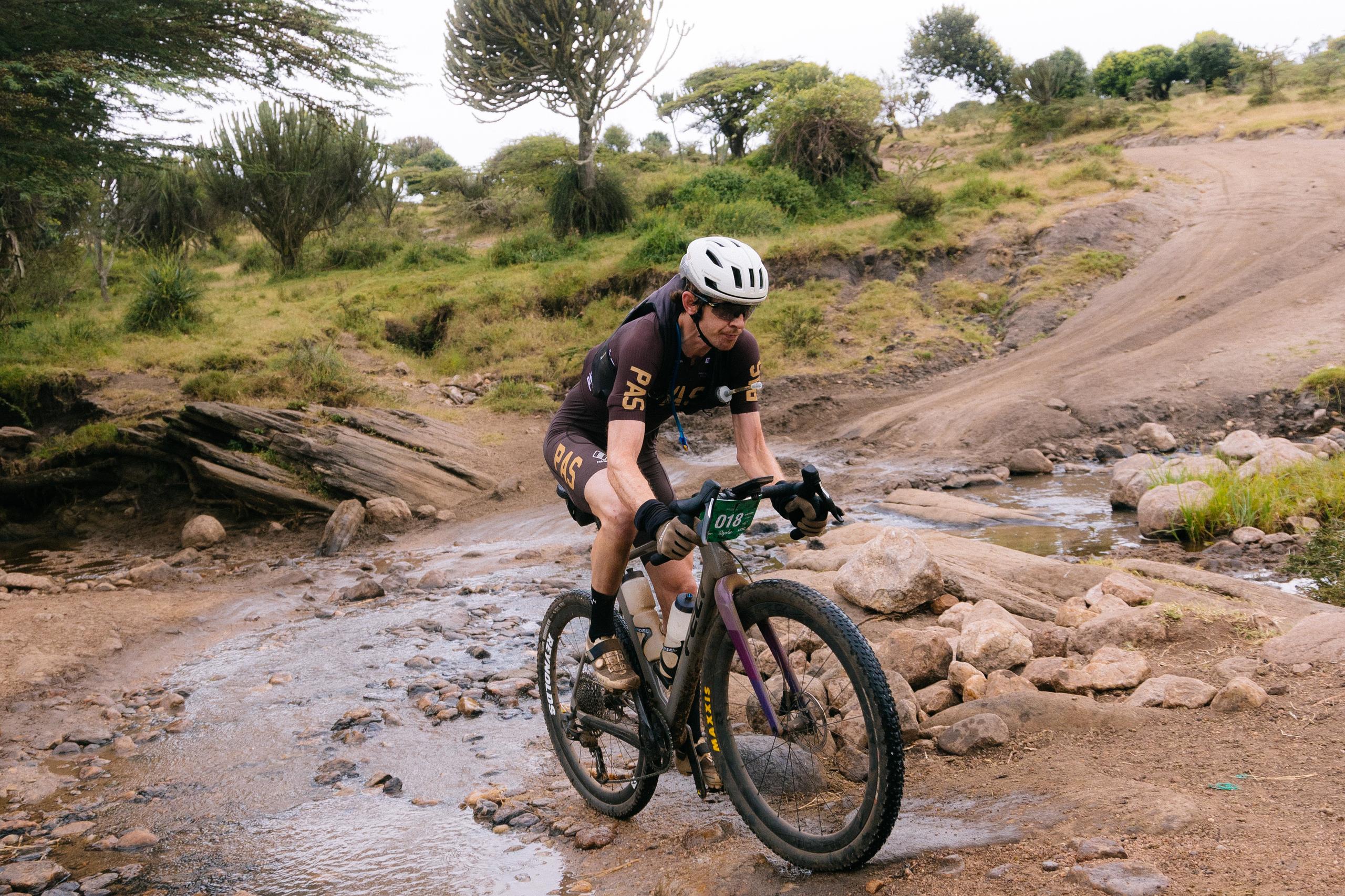 Model wearing black gravel cycling kit with yellow lettering racing mountain bike through rocky terrain