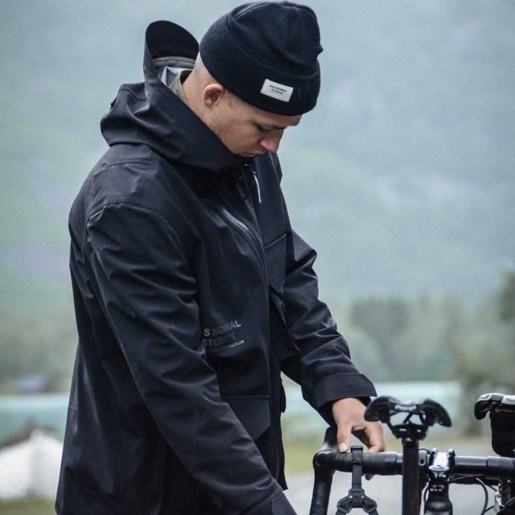 Model wearing black hooded jacket and beanie adjusting bicycle handlebars outdoors