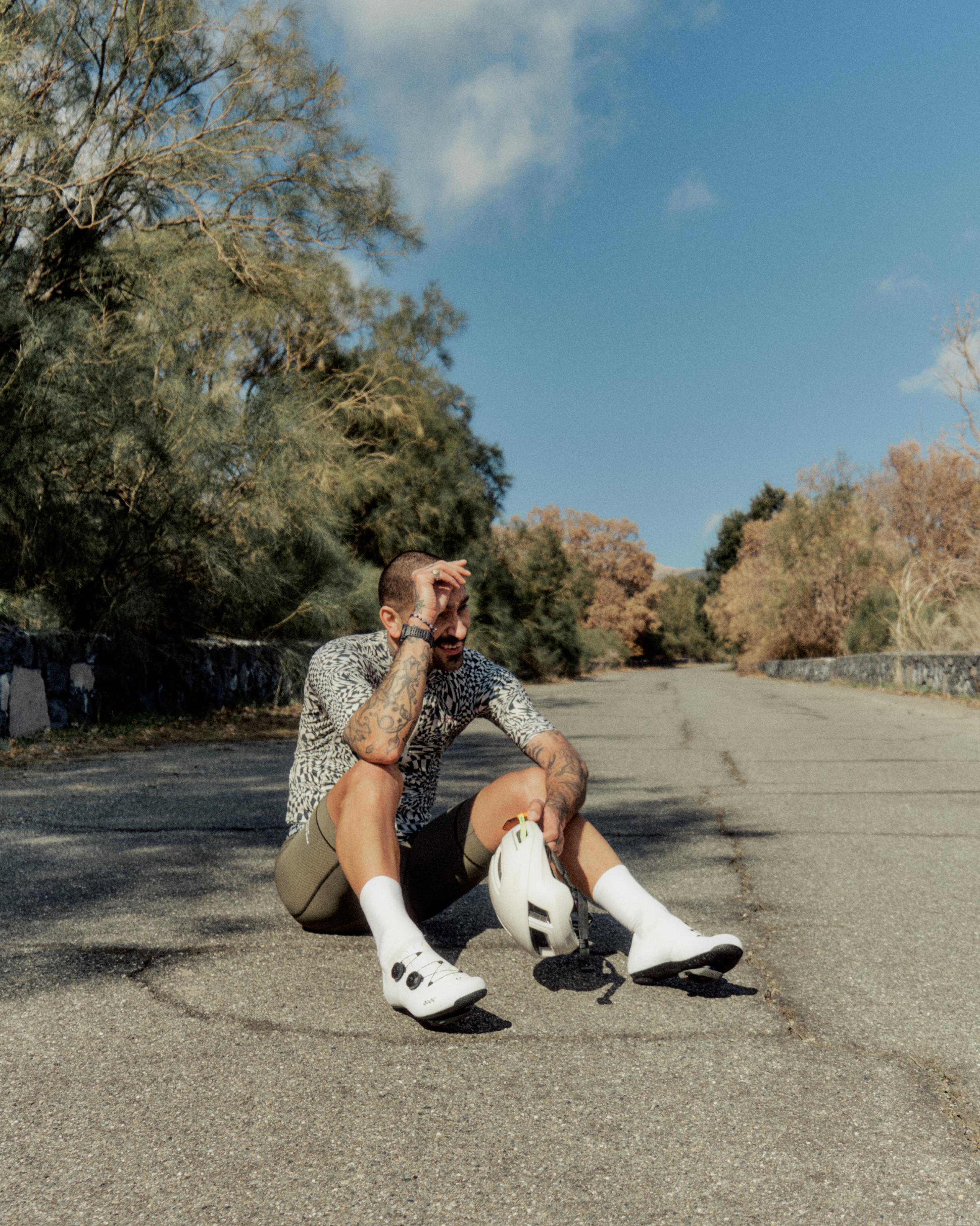 Model wearing printed jersey and black shorts with white cycling shoes, sitting on rural road in Sicily