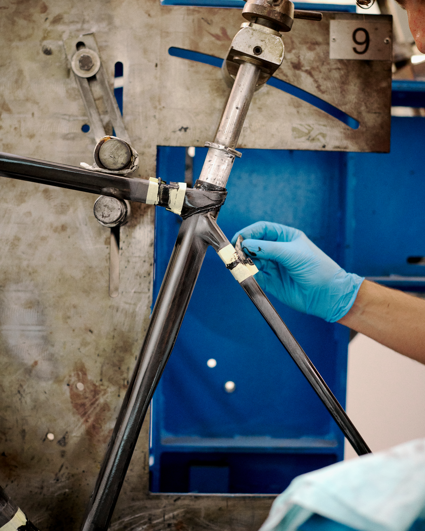 Bicycle frame being assembled in workshop with gloved hands adjusting components on blue workstation