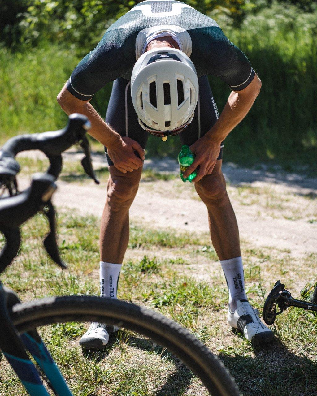 Model wearing black cycling jersey and shorts taking hydration break on gravel path with bicycle