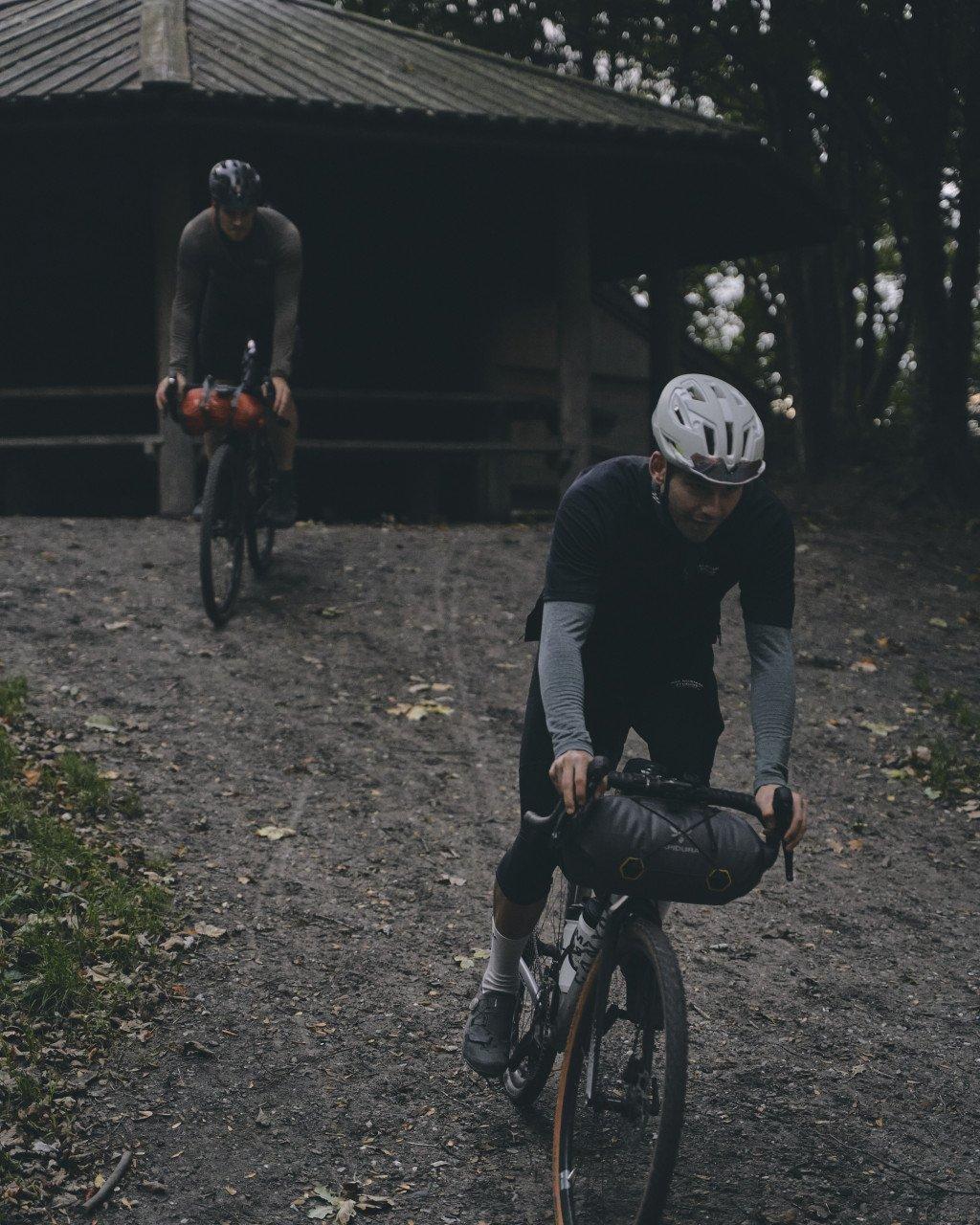 Two cyclists in dark cycling apparel preparing bikes for gravel ride at outdoor shelter