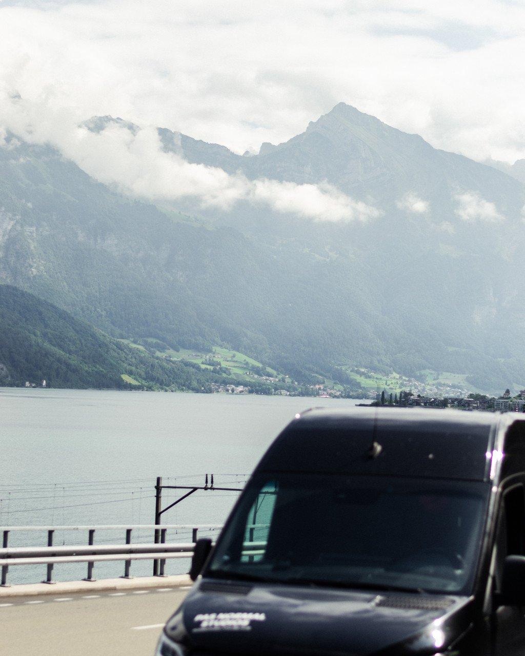 Dark van on lakeside road with misty mountains and cloudy peaks in background