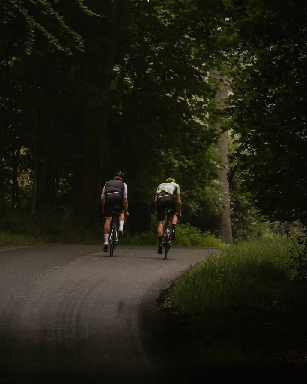 Two cyclists riding through tree-lined road in cycling jerseys and shorts