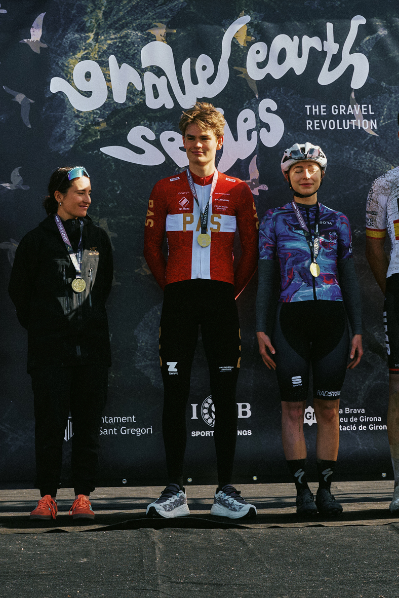 Three cyclists on podium with medals, center rider wearing red jersey, right rider in blue patterned jersey