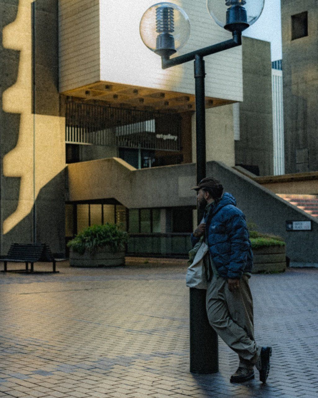 Model wearing blue jacket and grey trousers leaning against lamp post in urban plaza