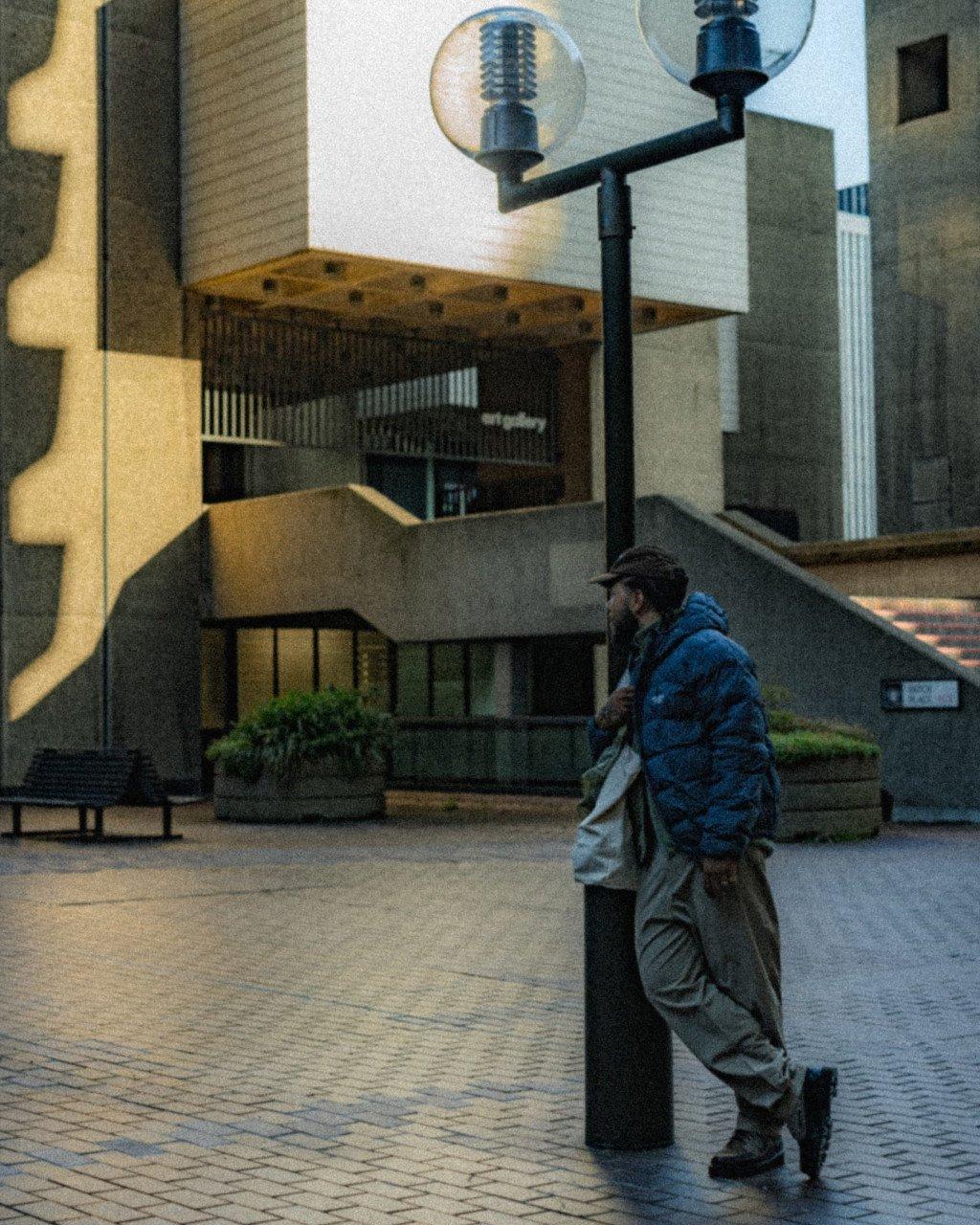 Model wearing blue jacket and grey trousers leaning against lamp post in urban plaza