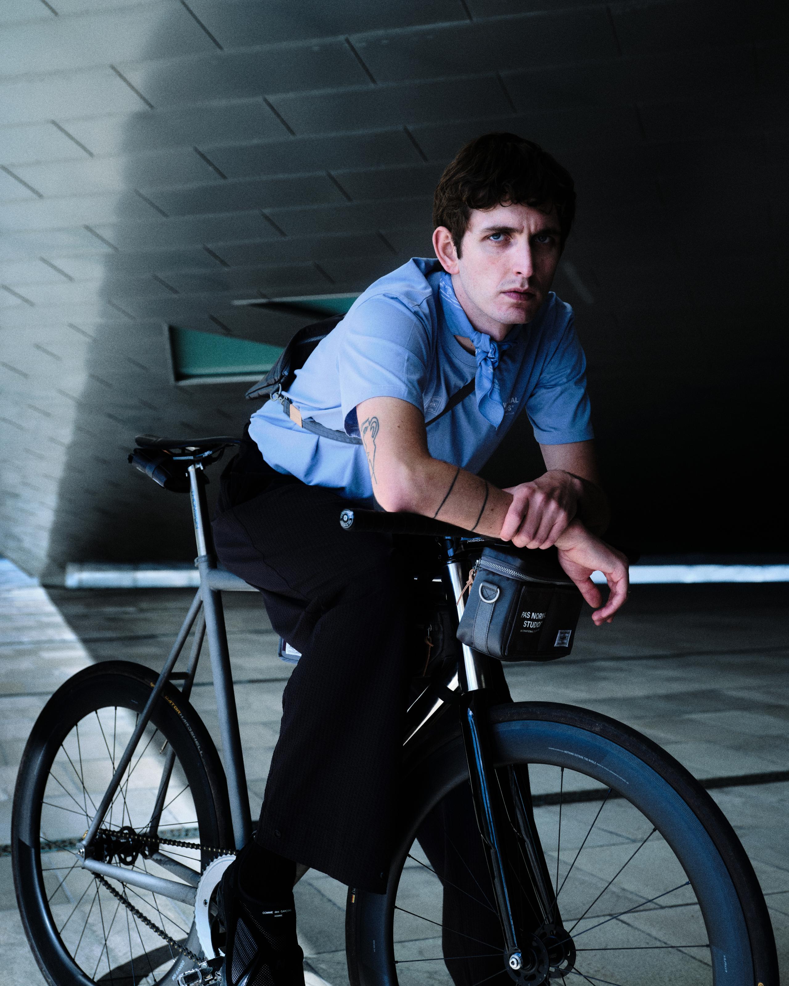 Model wearing blue cycling jersey with black shorts and frame bag, standing with bike in urban tunnel
