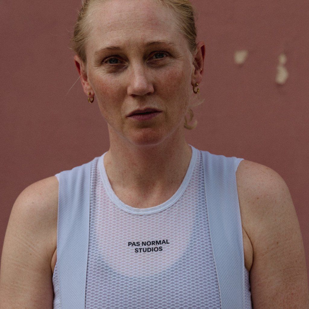Model wearing white sleeveless base layer with mesh panel and chest branding on track