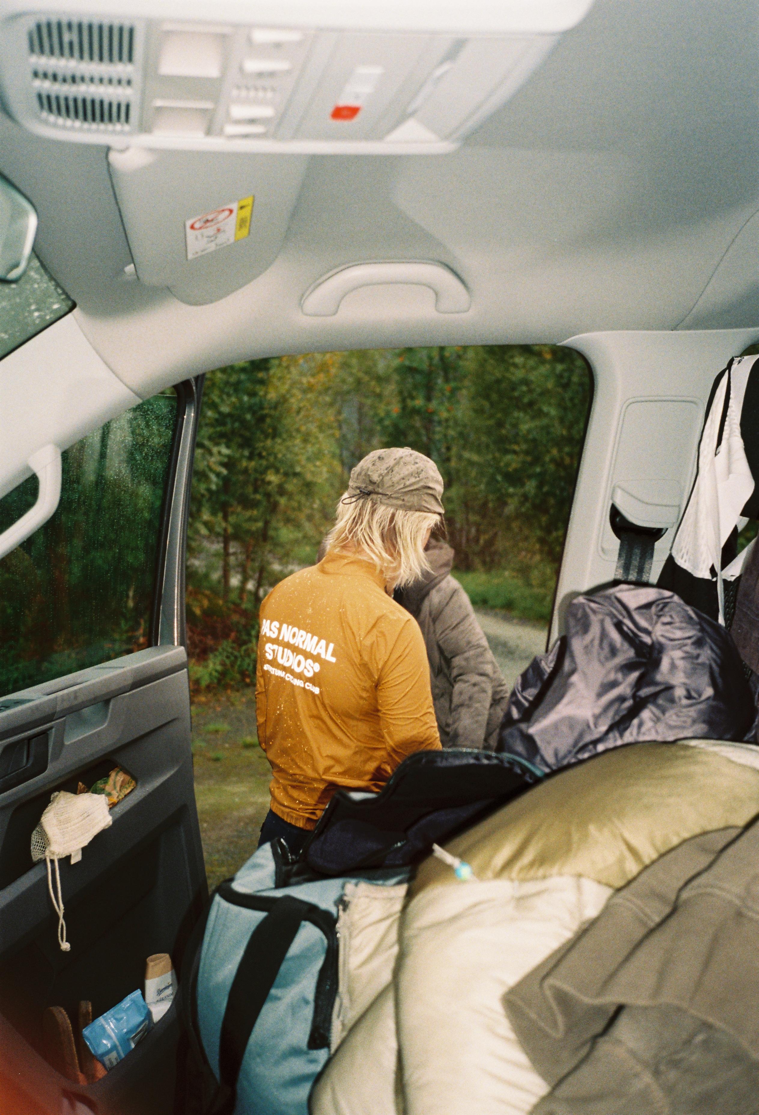 Model wearing orange insulated jacket with white back lettering sitting in van with outdoor gear