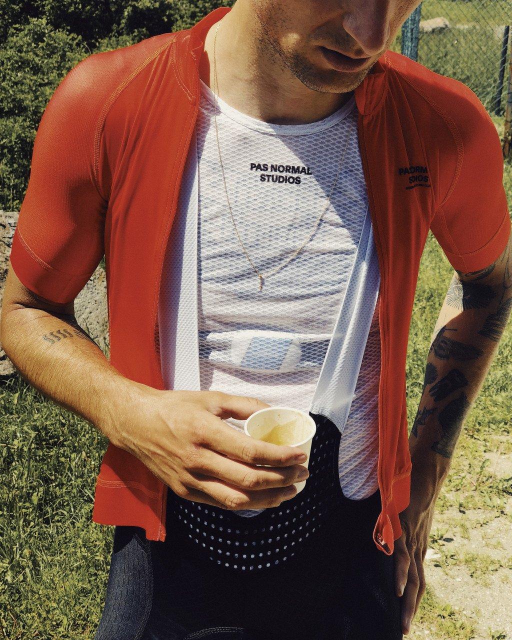 Model wearing red Mechanism Jersey over white mesh base layer with chest branding, holding drink outdoors