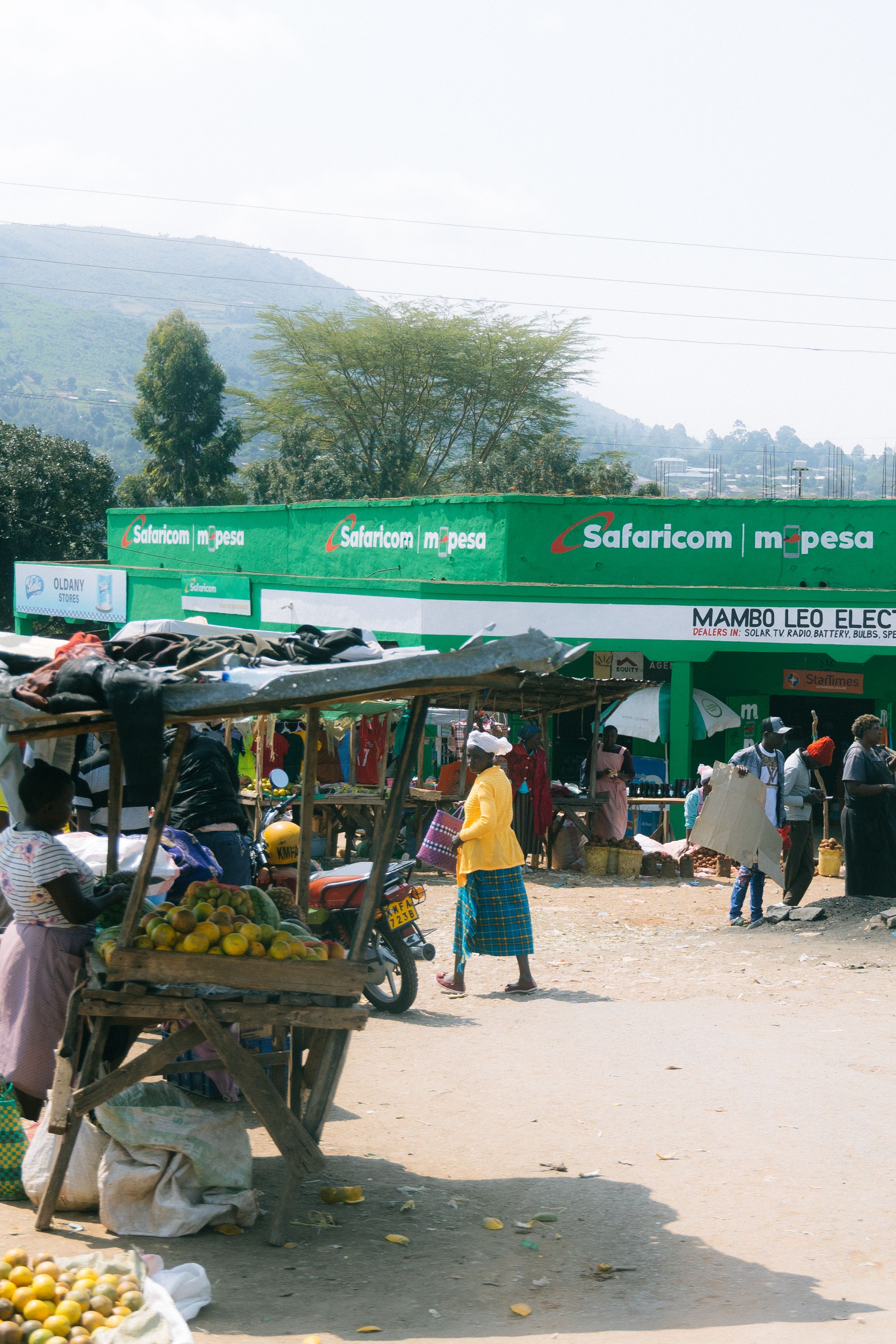 Local market scene with fruit vendors and green Safaricom signage in hilly landscape