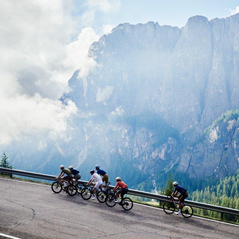 Group of cyclists riding on mountain road with dramatic alpine peaks and clouds in background