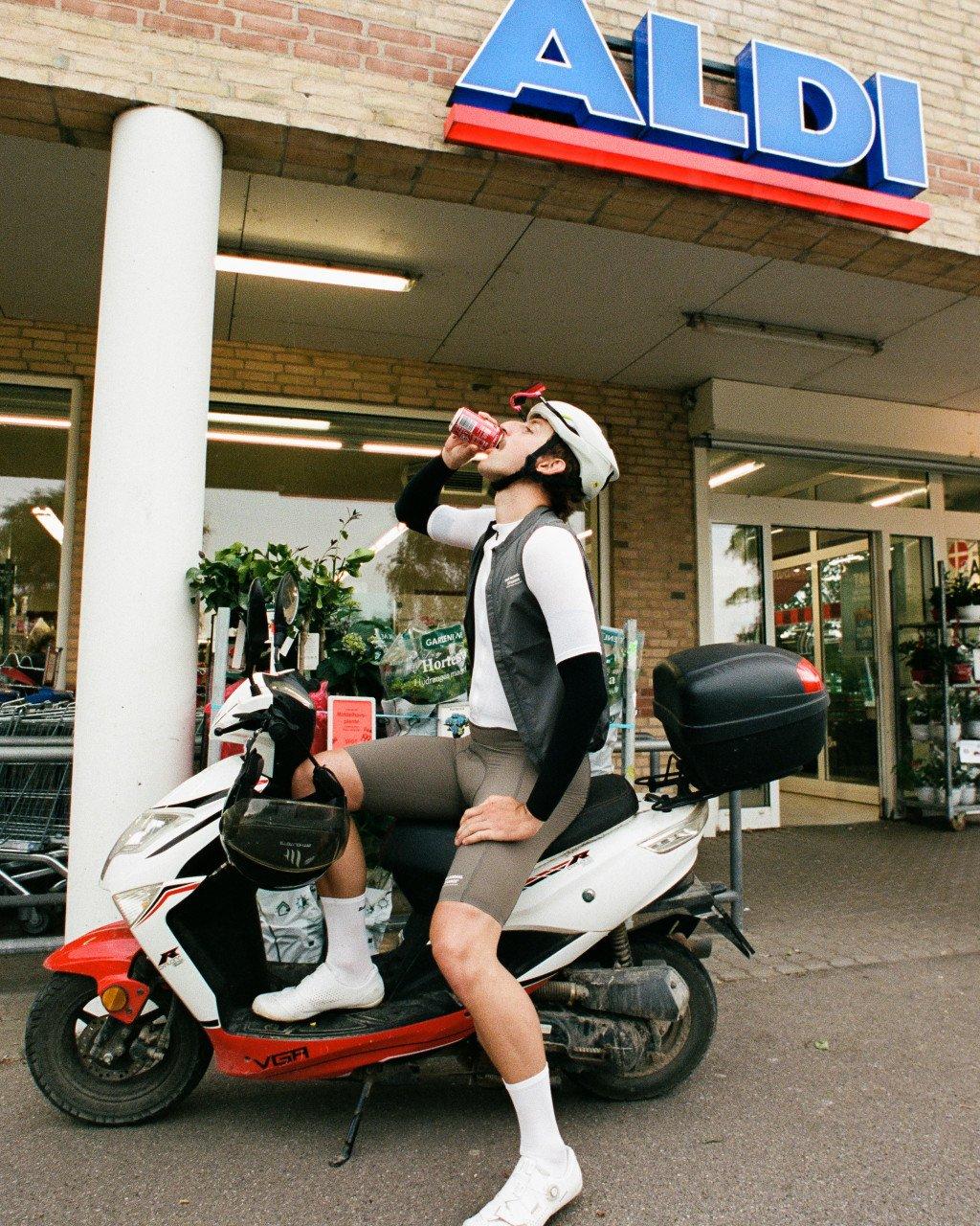 Model wearing black and white cycling kit drinking from bottle while sitting on scooter outside ALDI store