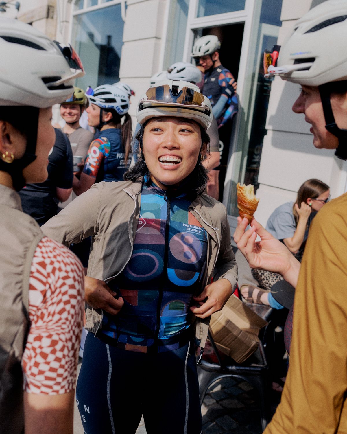 Female cyclist wearing multicolored graphic jersey and beige jacket in group setting with helmets