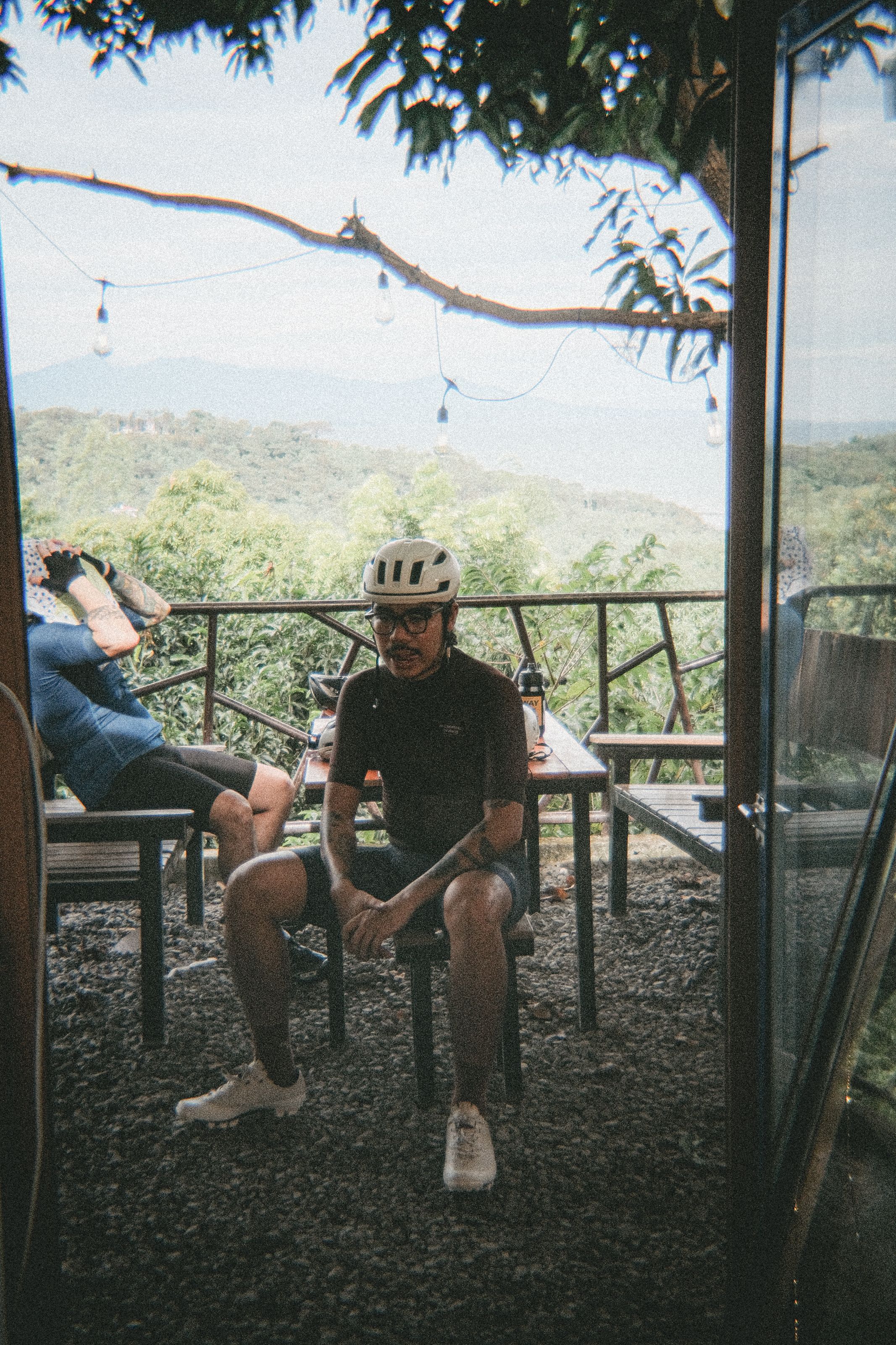 Two cyclists relaxing on terrace overlooking mountain landscape, one wearing dark jersey and helmet