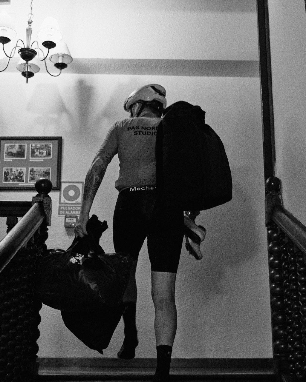 Model wearing cycling jersey with back branding and black shorts, carrying jacket indoors in black and white
