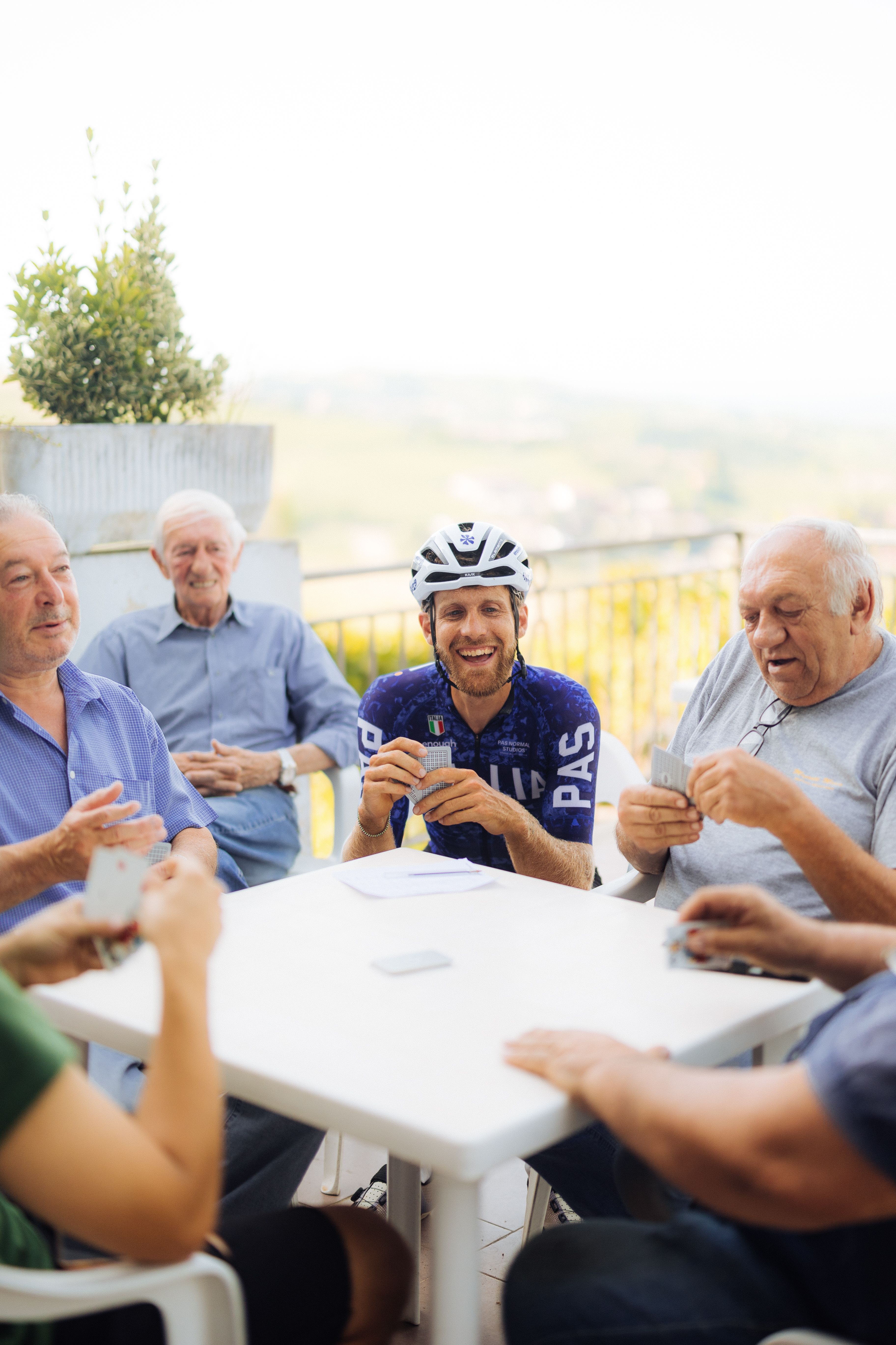 Cyclist in blue jersey with branding playing cards with group of men at outdoor table
