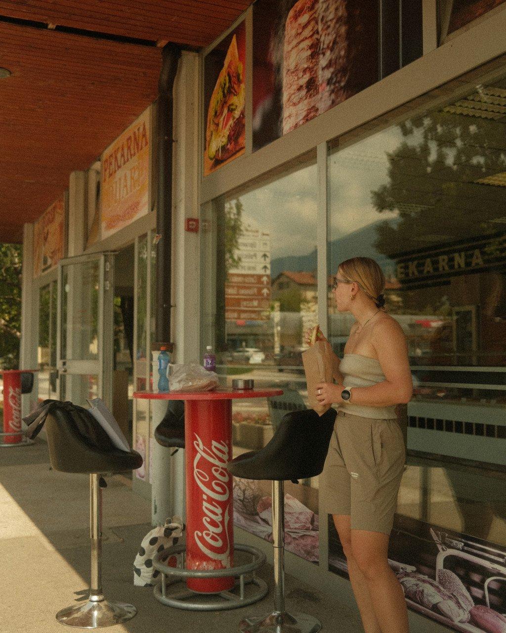 Model wearing beige strapless top and shorts at European cafe with Coca-Cola table