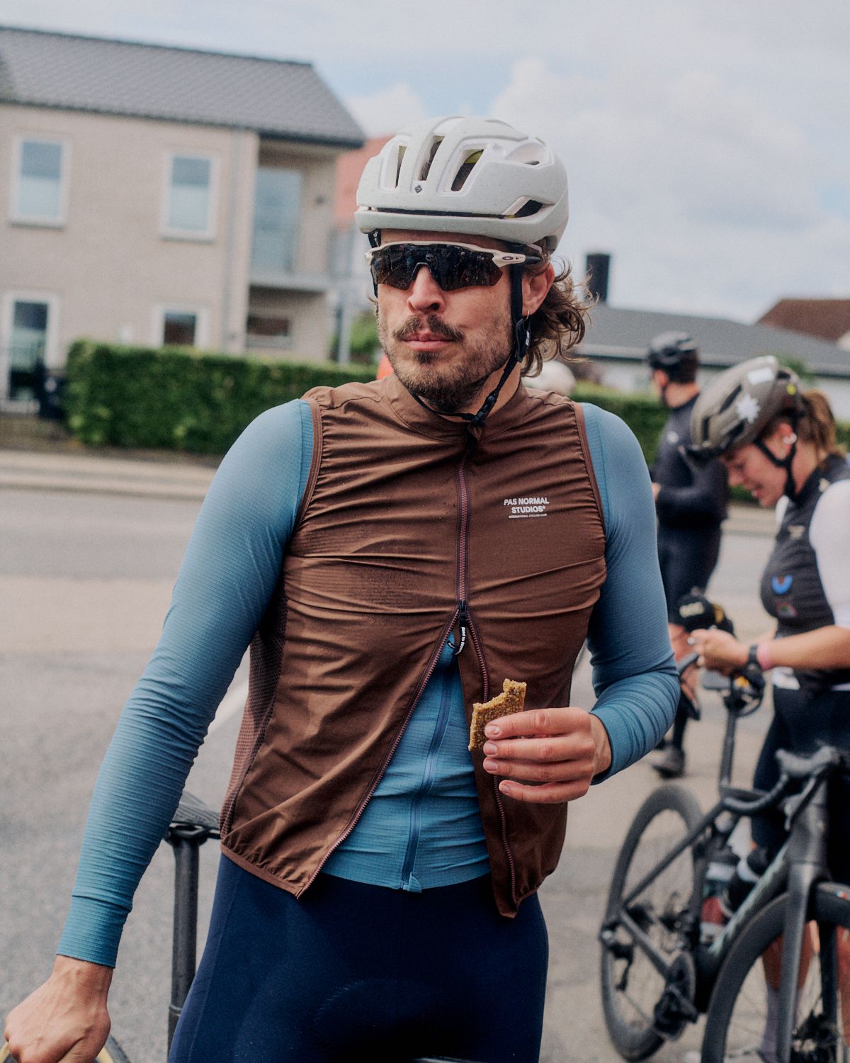 Model wearing brown Insulation Vest with blue long sleeve jersey underneath, holding snack during cycling event