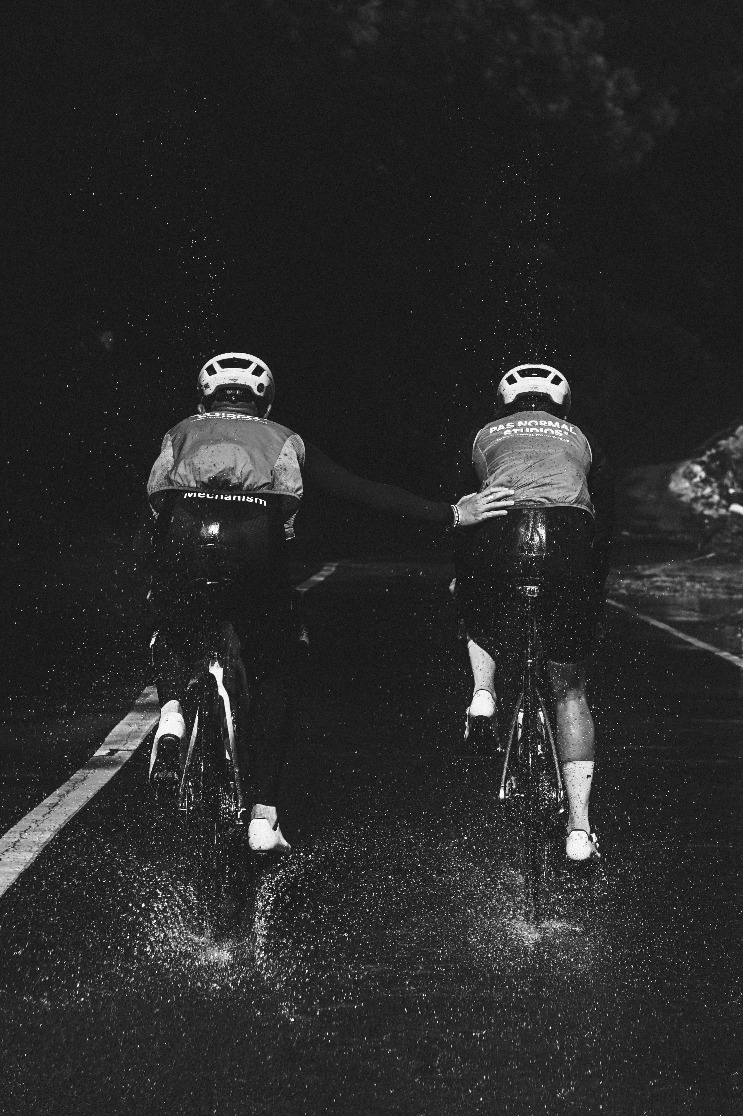Two cyclists in aerodynamic position on wet road in rain, wearing cycling jerseys with back branding