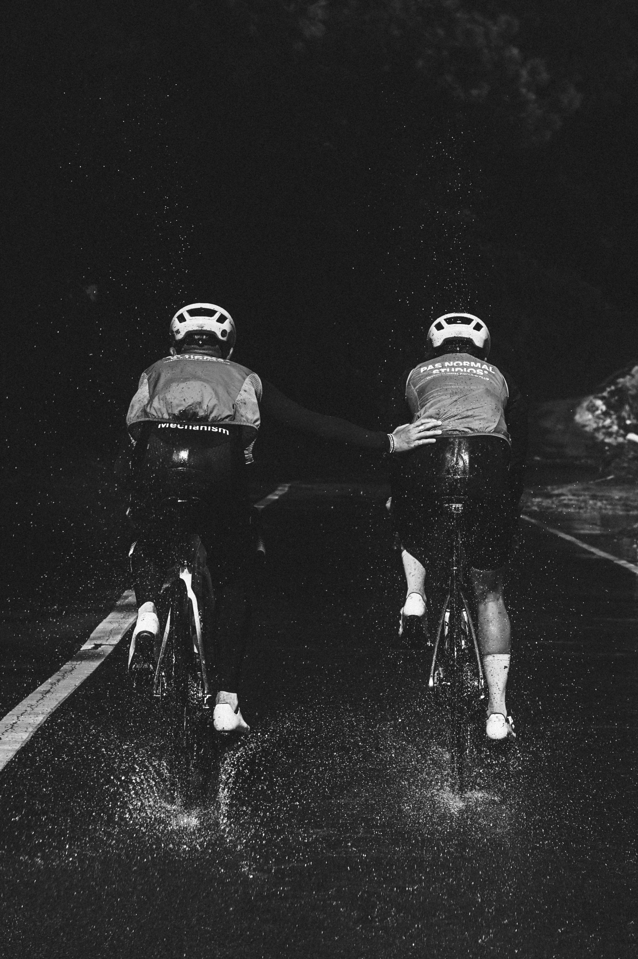 Two cyclists in aerodynamic position on wet road in rain, wearing cycling jerseys with back branding