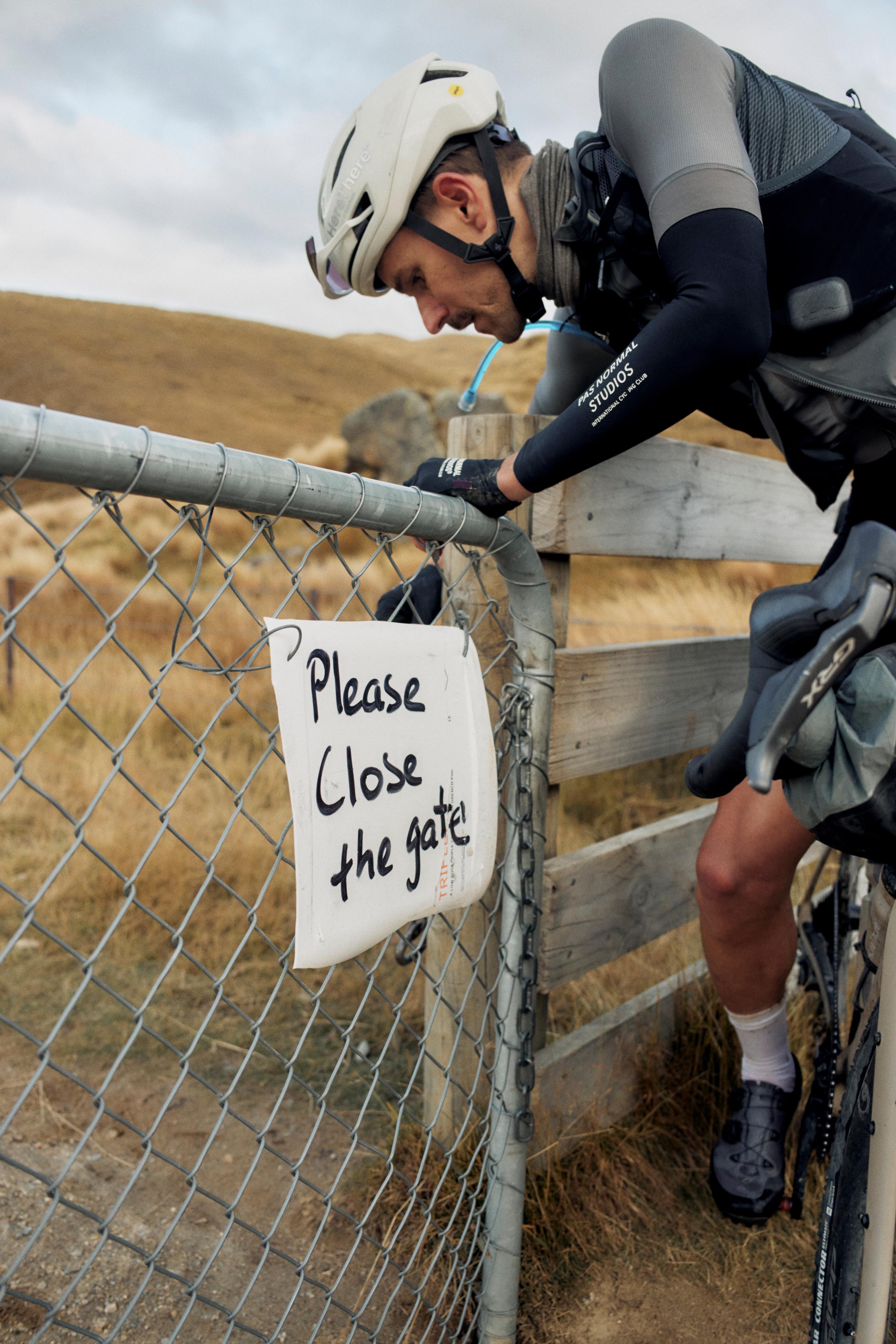 Model wearing navy long sleeve jersey with branding, time trial helmet, and hydration pack at rural gate