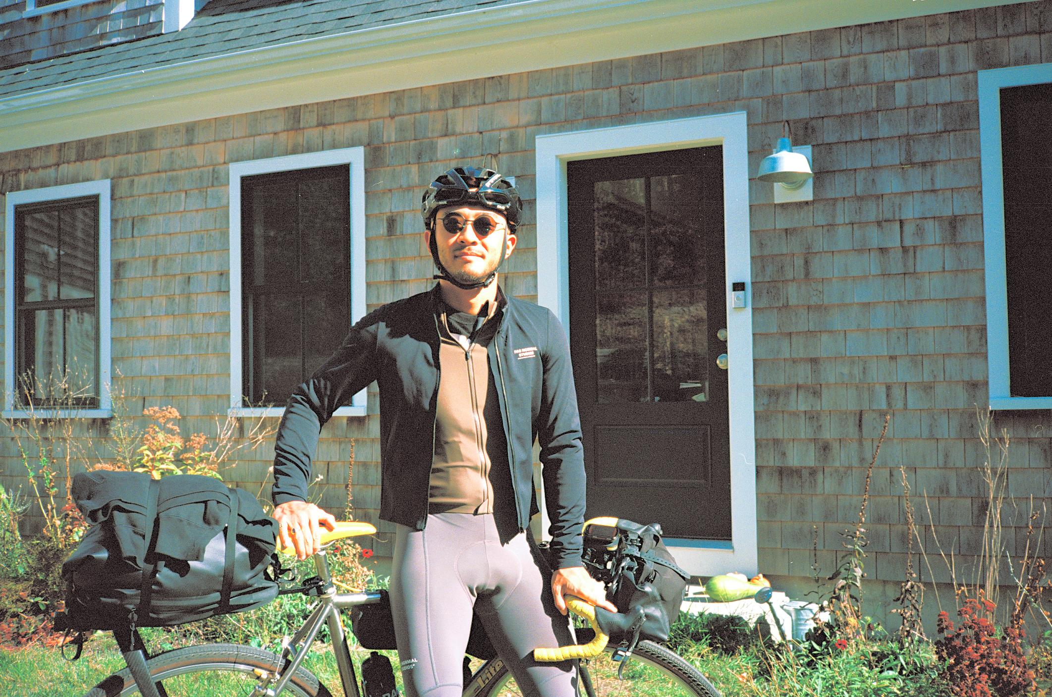 Model wearing dark jacket over cycling jersey with grey bib shorts, helmet and sunglasses, standing with bicycle