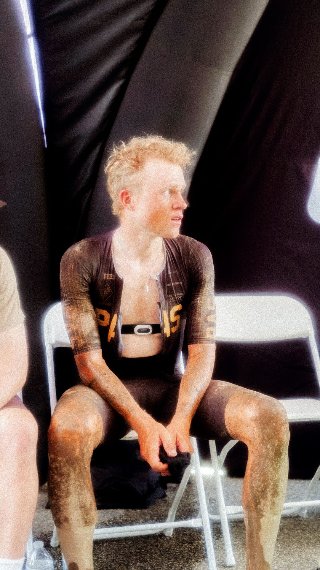 Model wearing dark brown long sleeve cycling jersey with orange lettering, sitting outdoors covered in dust