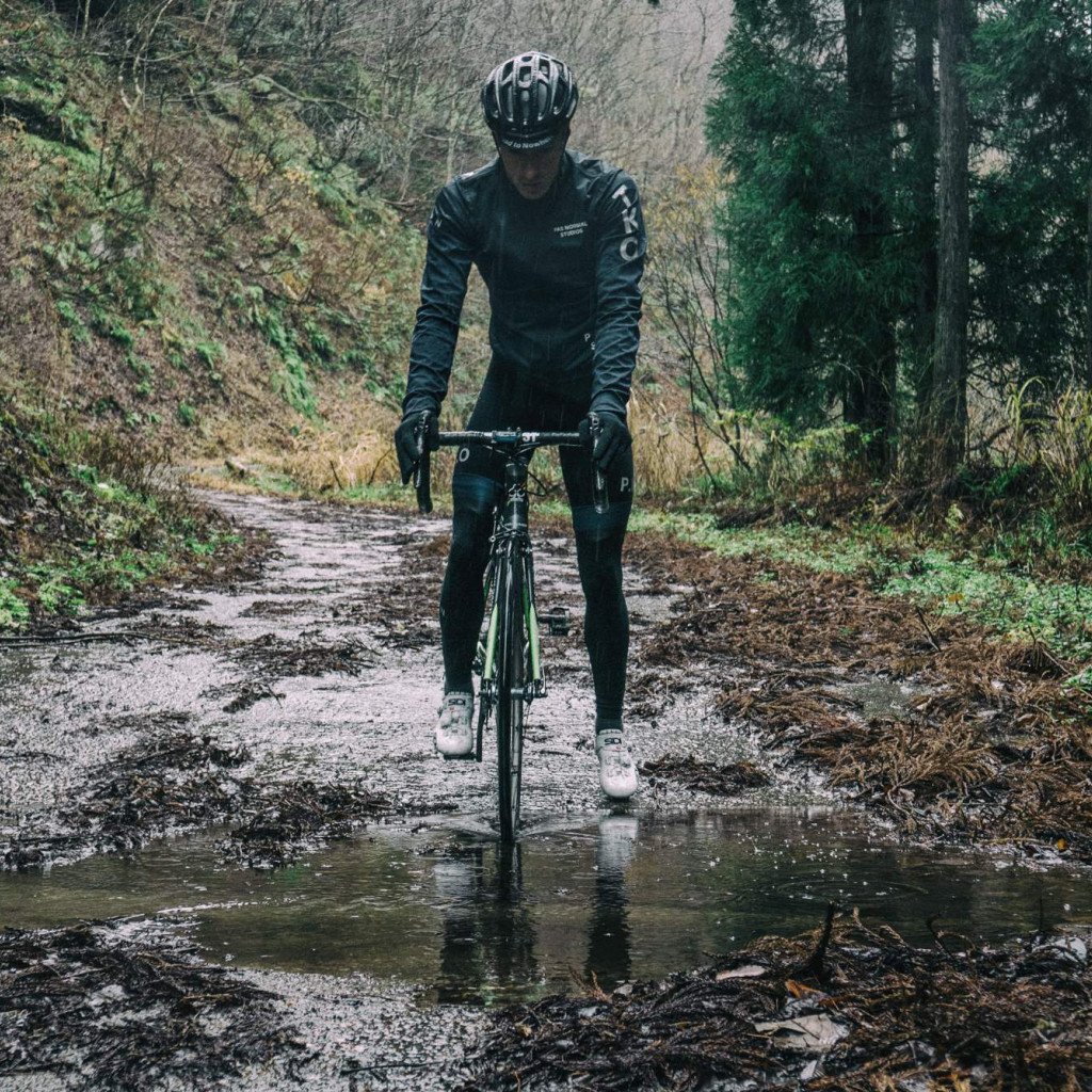 Model wearing black long-sleeve cycling jersey and tights on muddy forest trail with bike