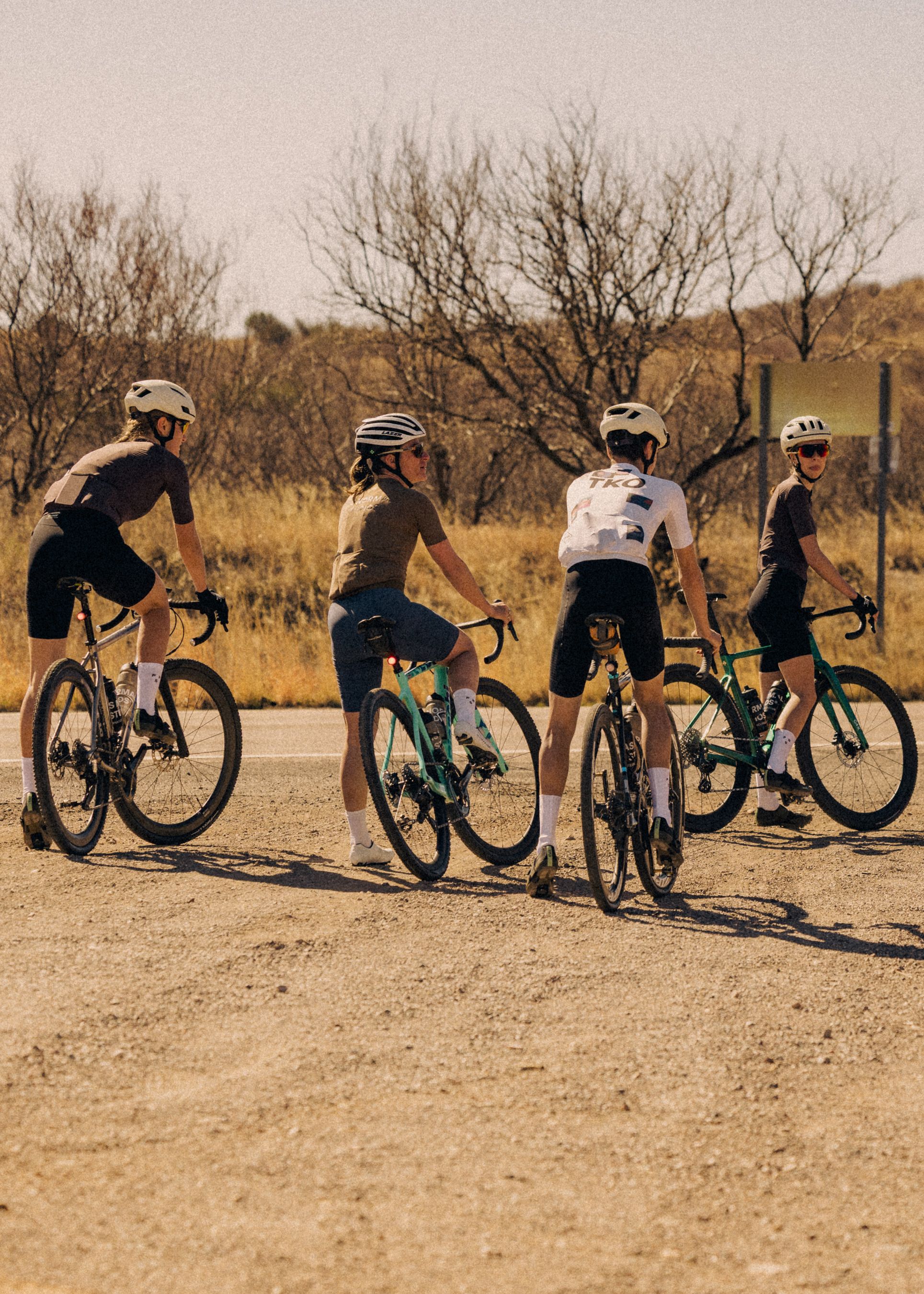 Four cyclists with road bikes on gravel terrain wearing mixed cycling apparel in desert landscape