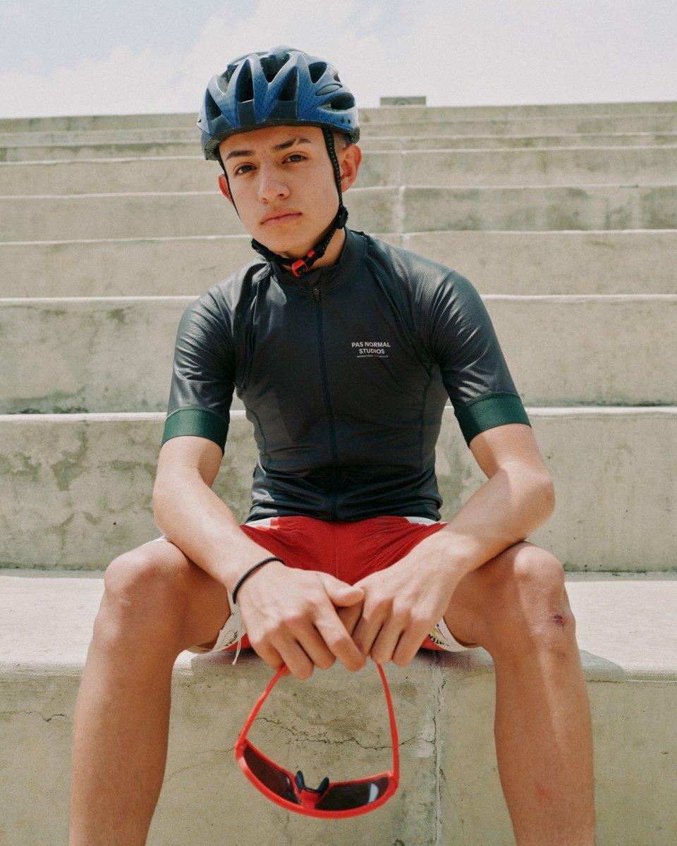Model wearing dark grey cycling jersey with green sleeve details and red shorts, sitting on concrete steps