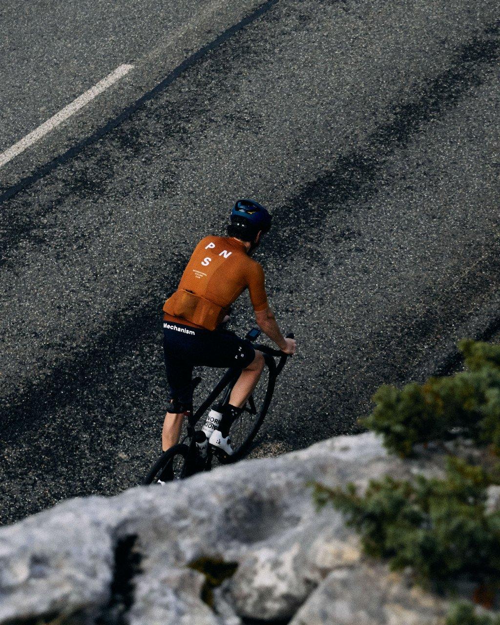 Model wearing orange Mechanism jersey and black shorts cycling on mountain road with blue helmet