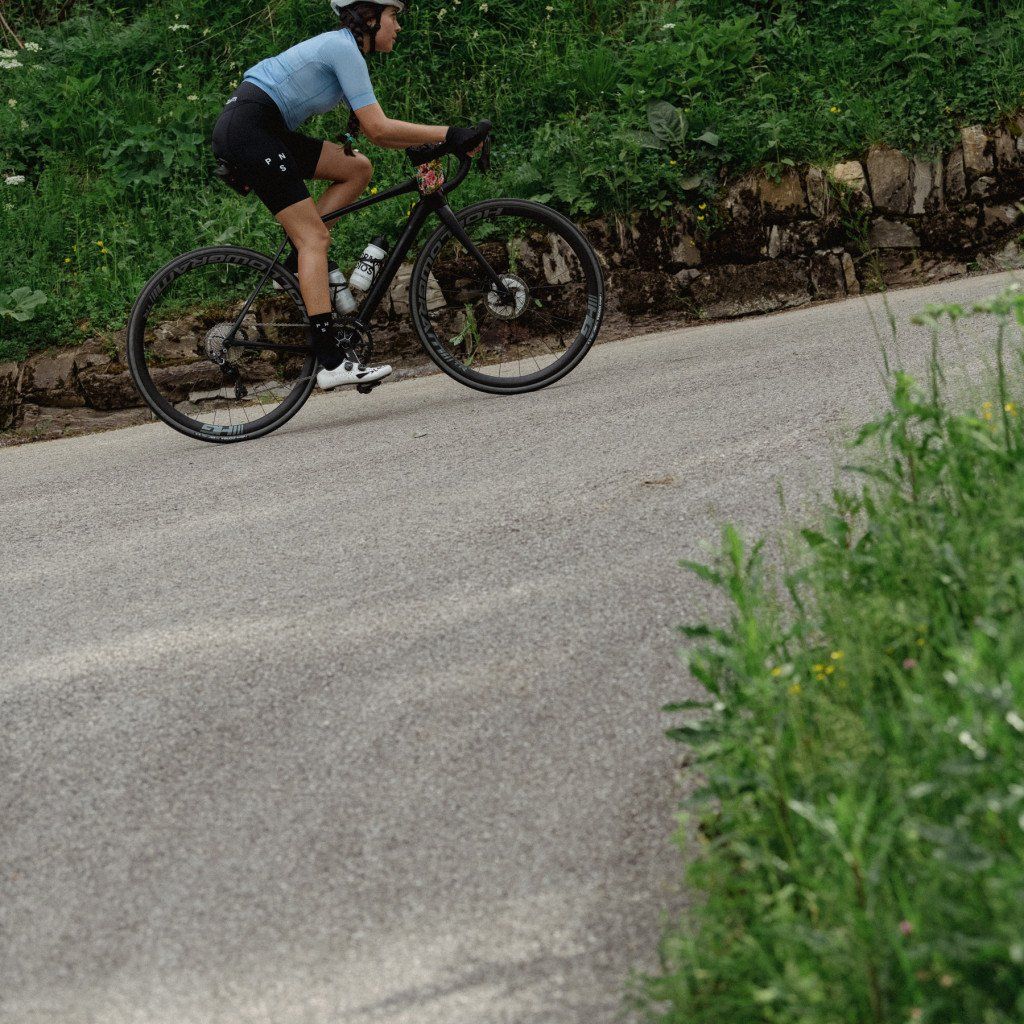 Model wearing light blue jersey and black cycling shorts on road bike in countryside setting