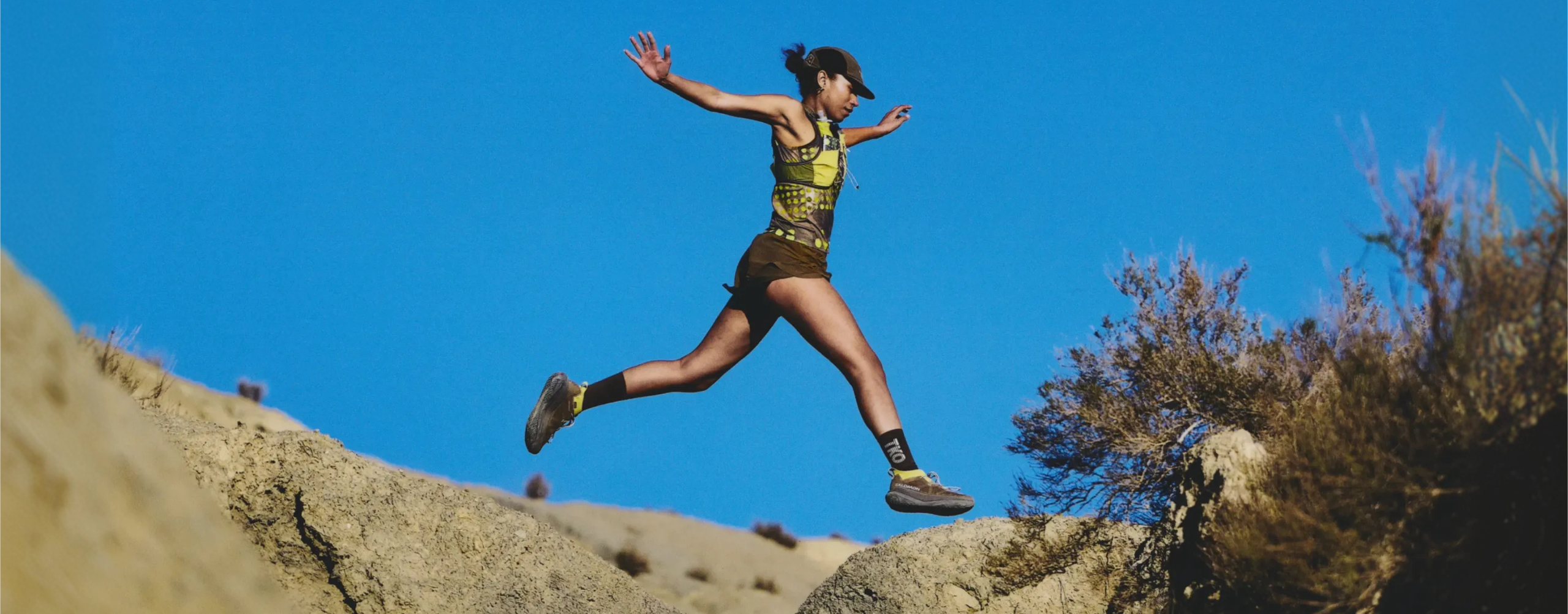 Model wearing yellow patterned top and brown shorts leaping across desert terrain against blue sky