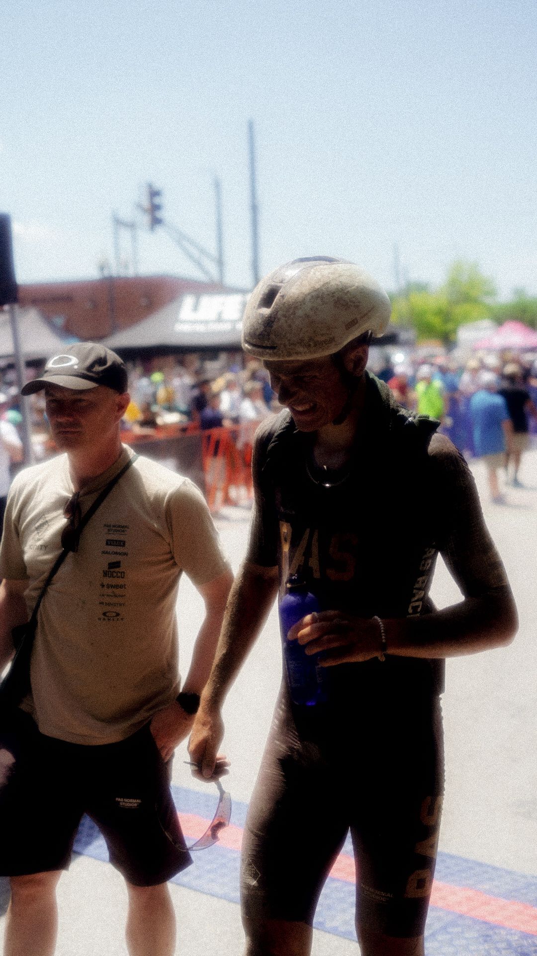 Two cyclists at gravel race event, one wearing white jersey with text, other in black kit with helmet