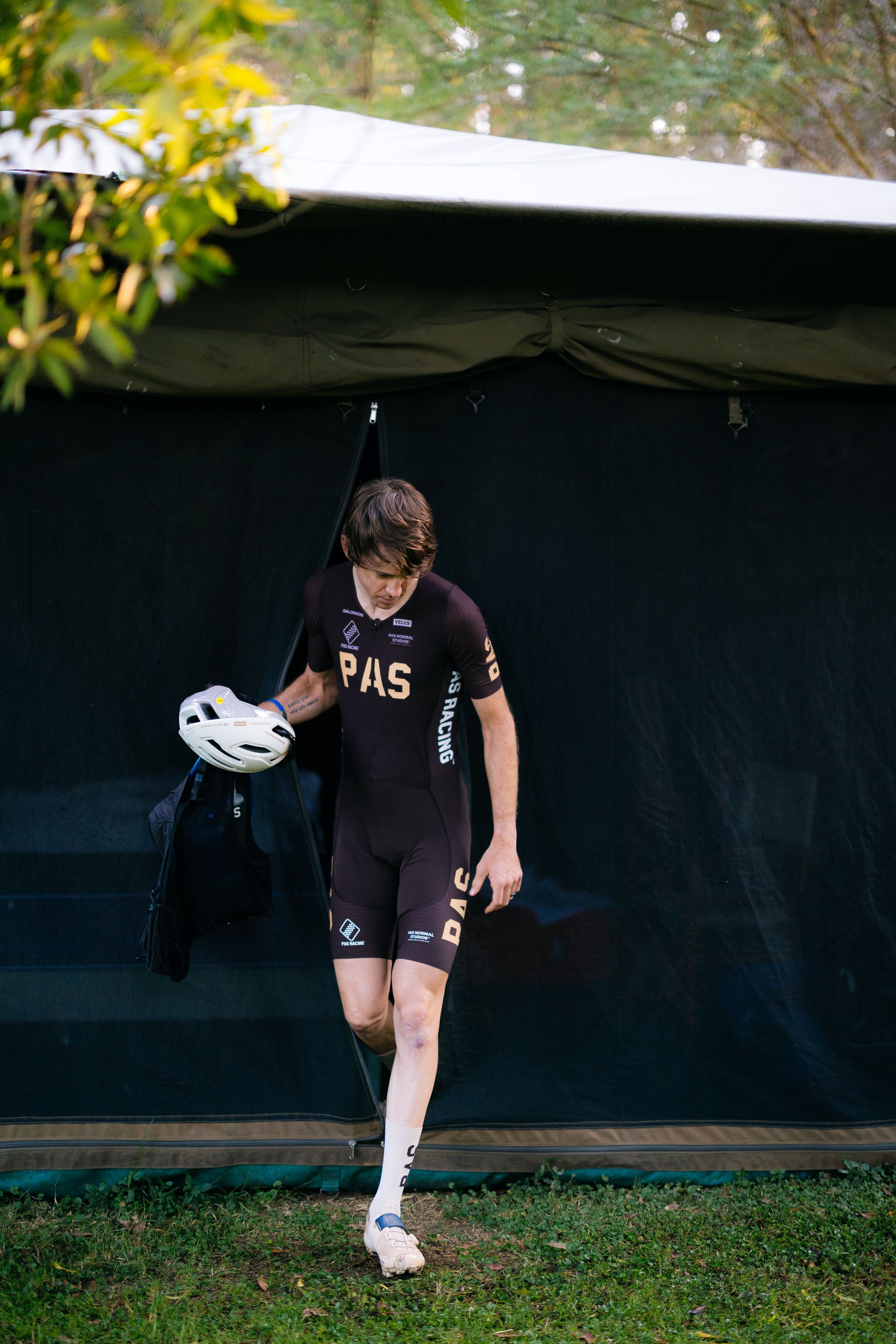 Model wearing dark purple PAS gravel kit with tan lettering, holding white helmet outside tent