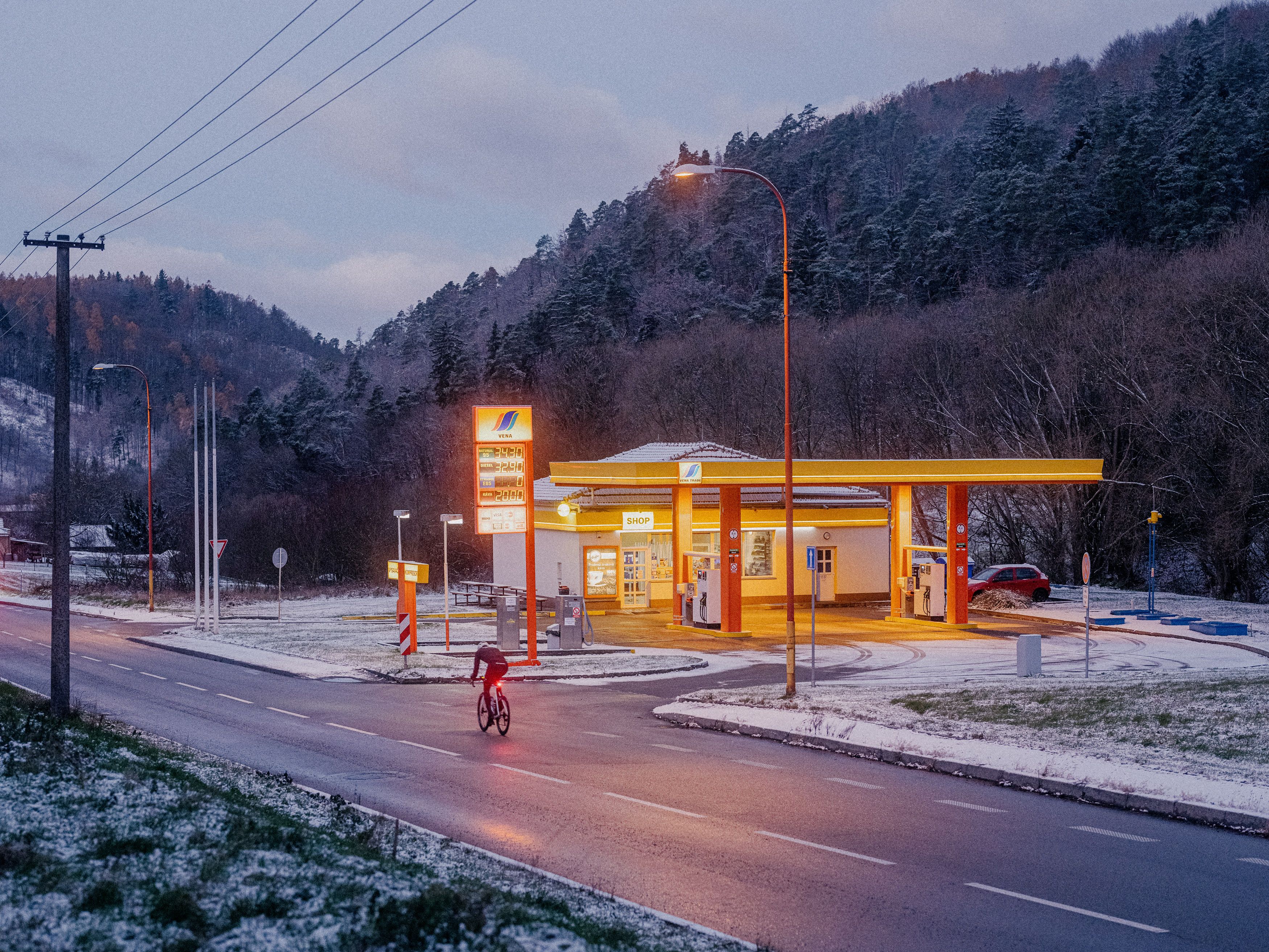Cyclist in red riding past illuminated gas station at dusk in mountainous winter landscape