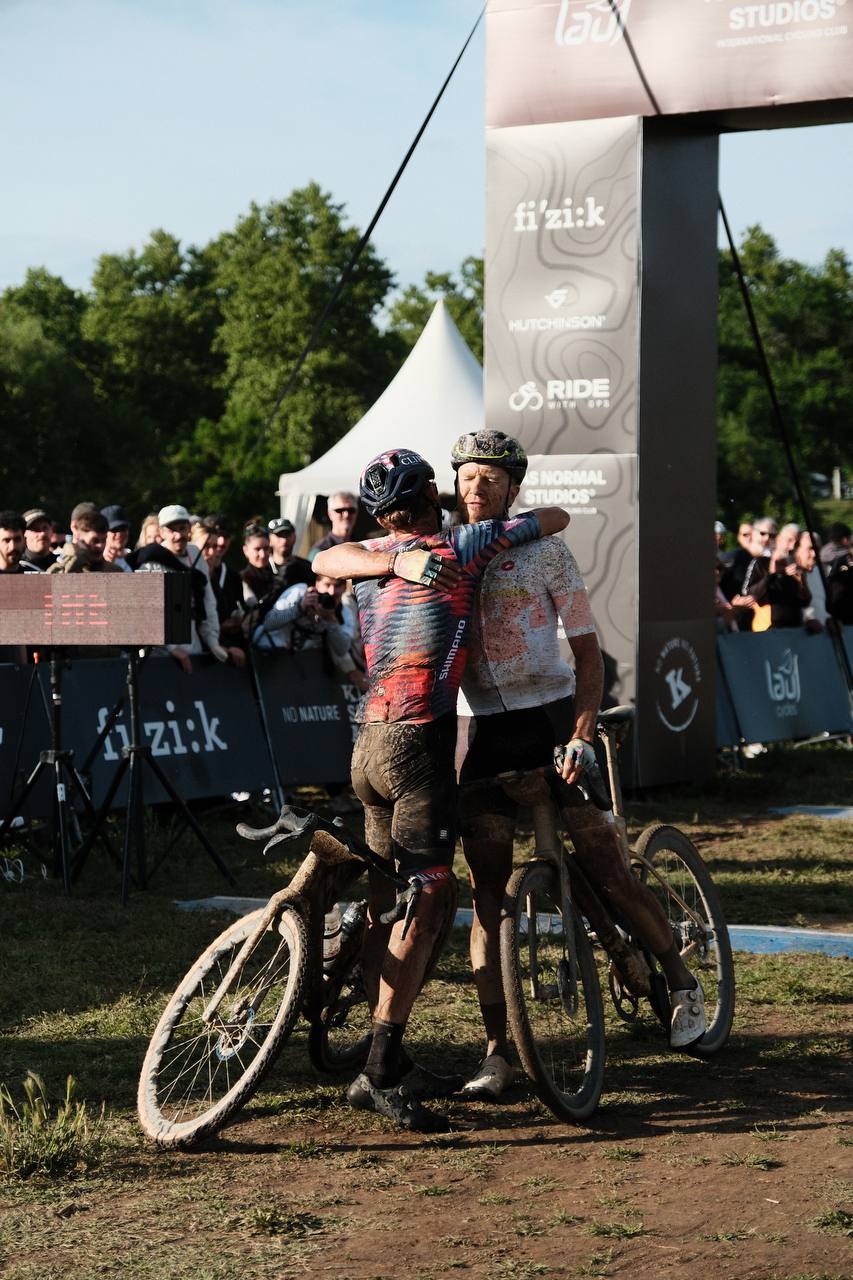 Two muddy cyclists embracing after finishing gravel race with bikes and crowd in background