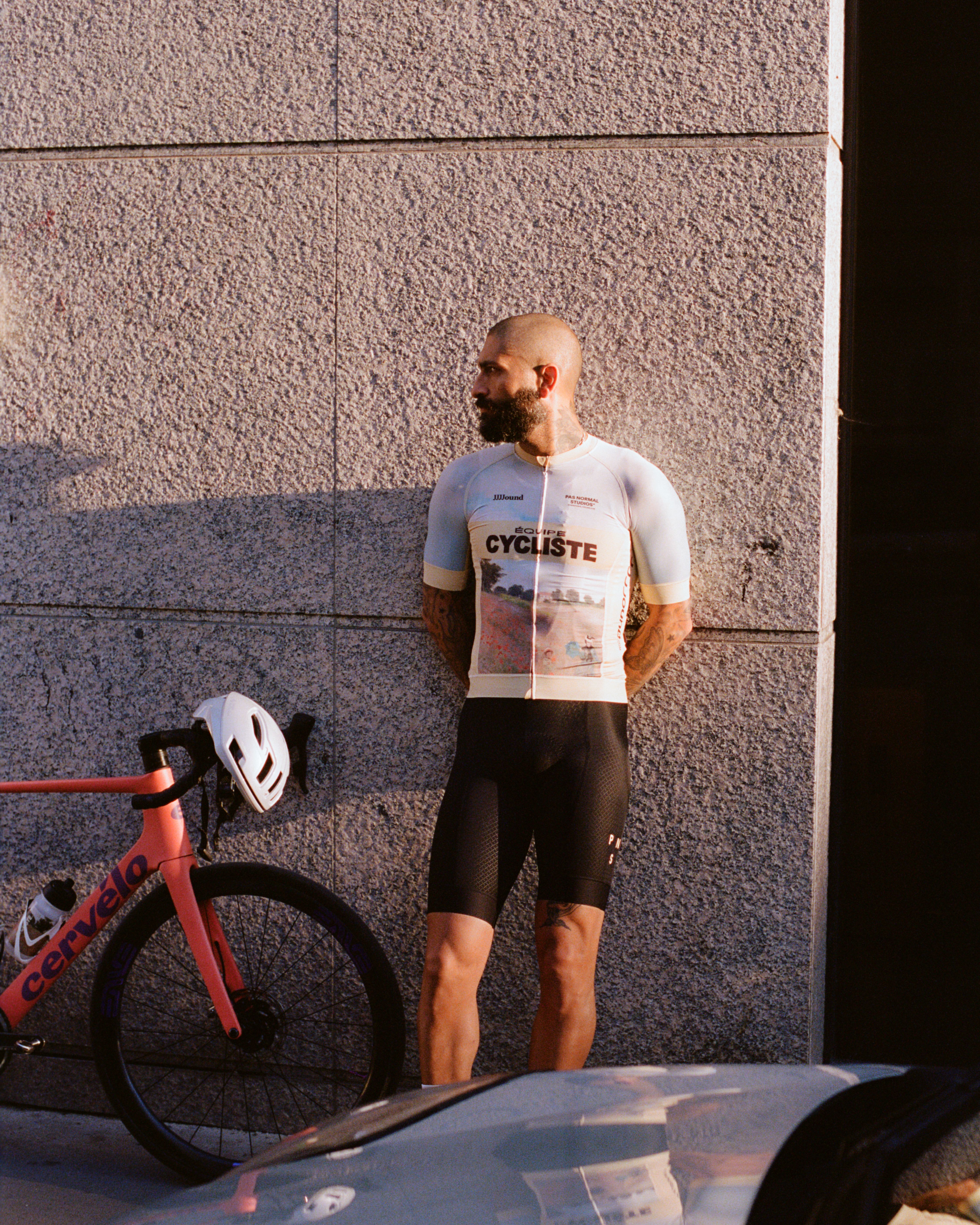 Model wearing white JJJJound collaboration jersey with graphic print and black shorts, standing beside bike