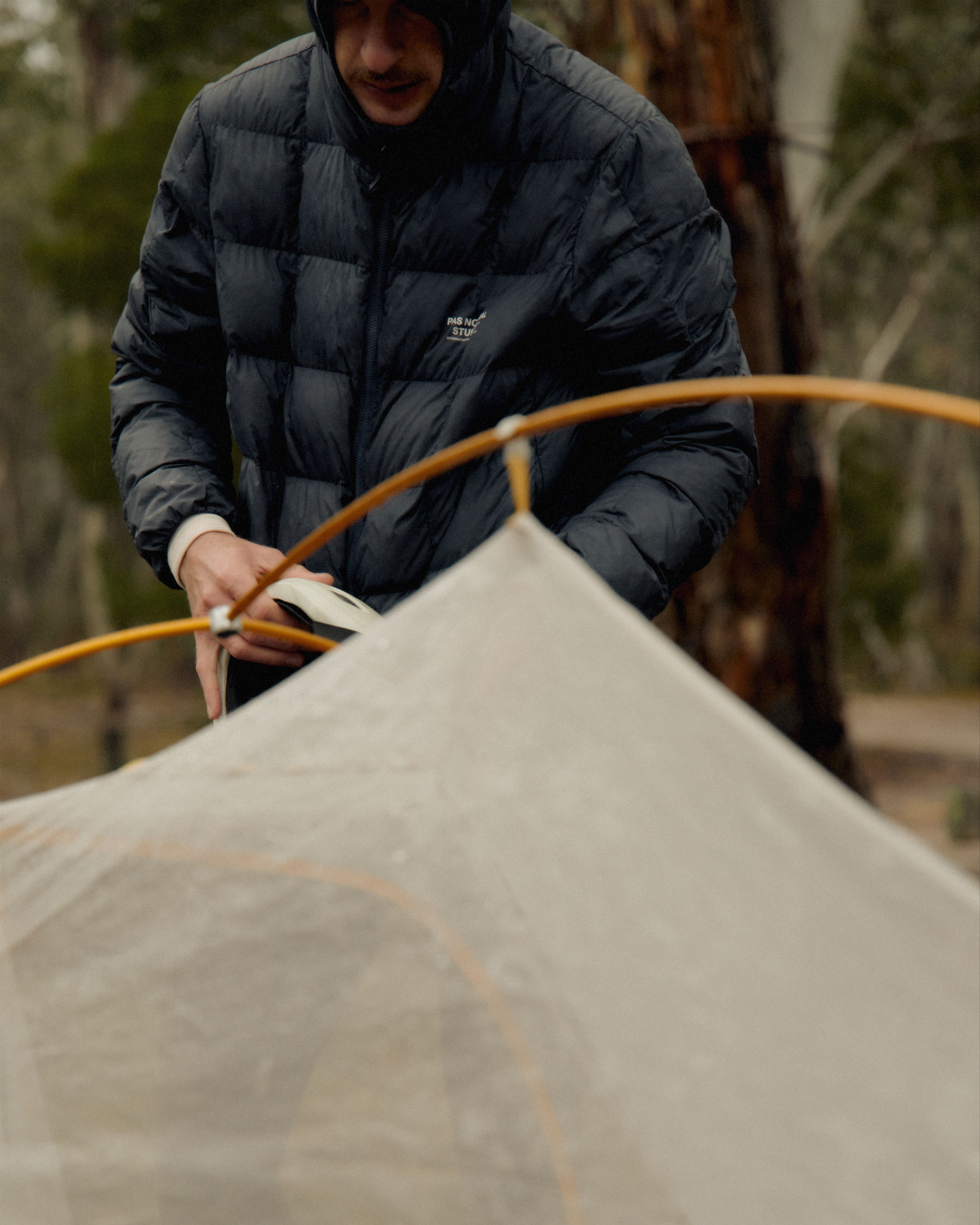 Model wearing dark grey quilted down jacket with logo while setting up tent outdoors