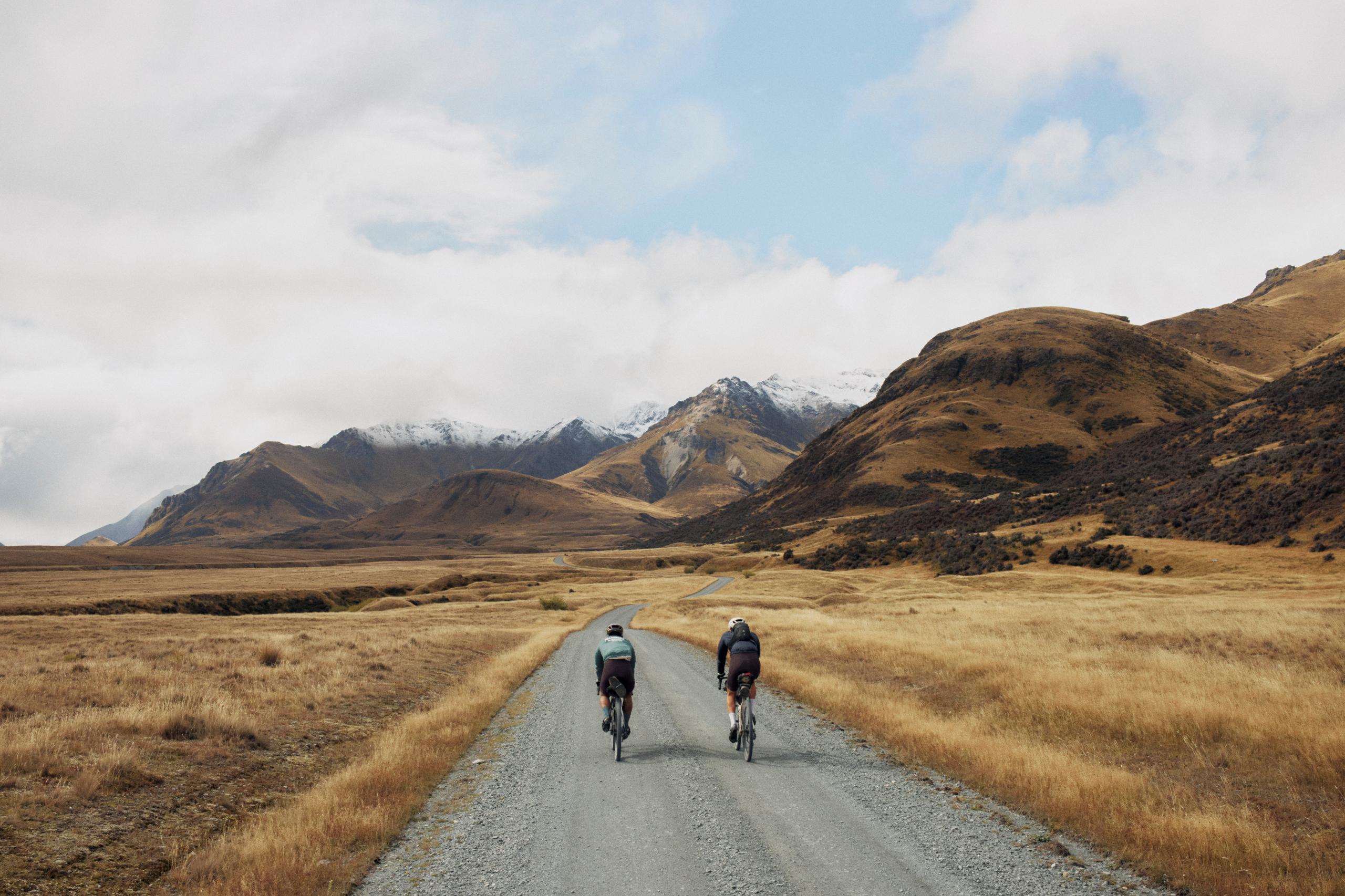 Two cyclists riding on gravel road through golden grassland with snow-capped mountains in background