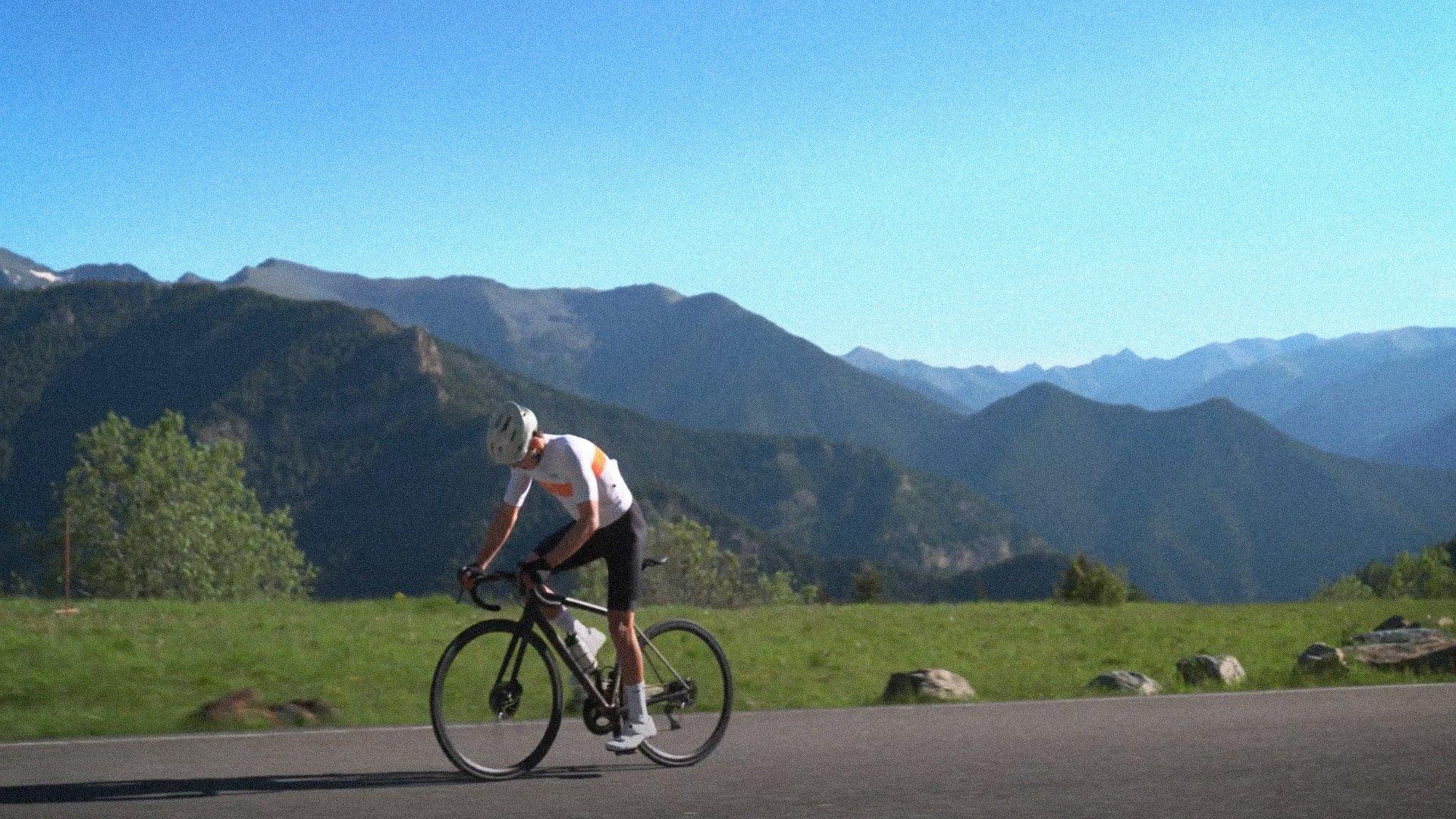 Model wearing white jersey with orange accents cycling on mountain road with alpine landscape