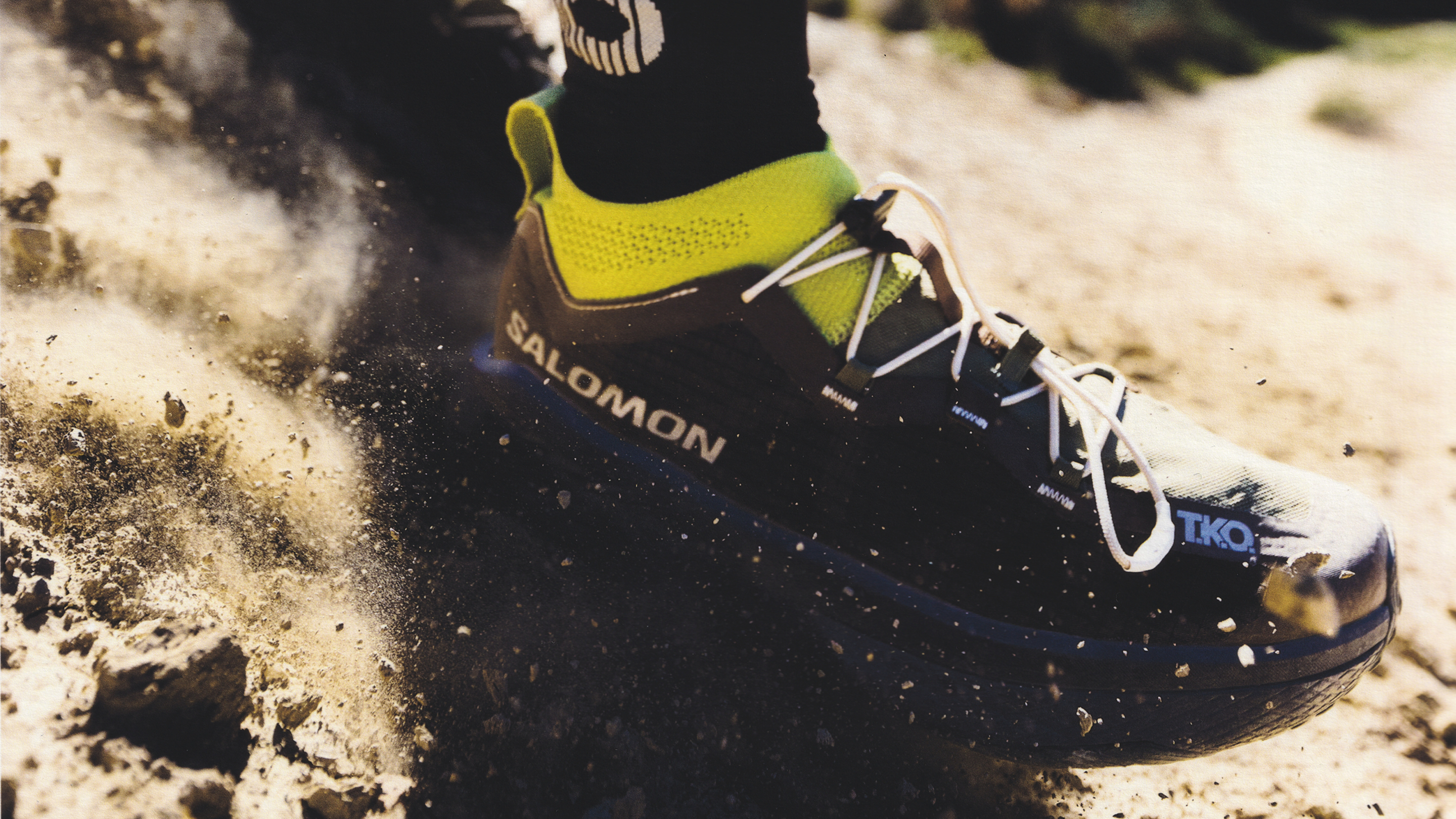 Black and green Salomon cycling shoe with white laces kicking up sand during outdoor activity