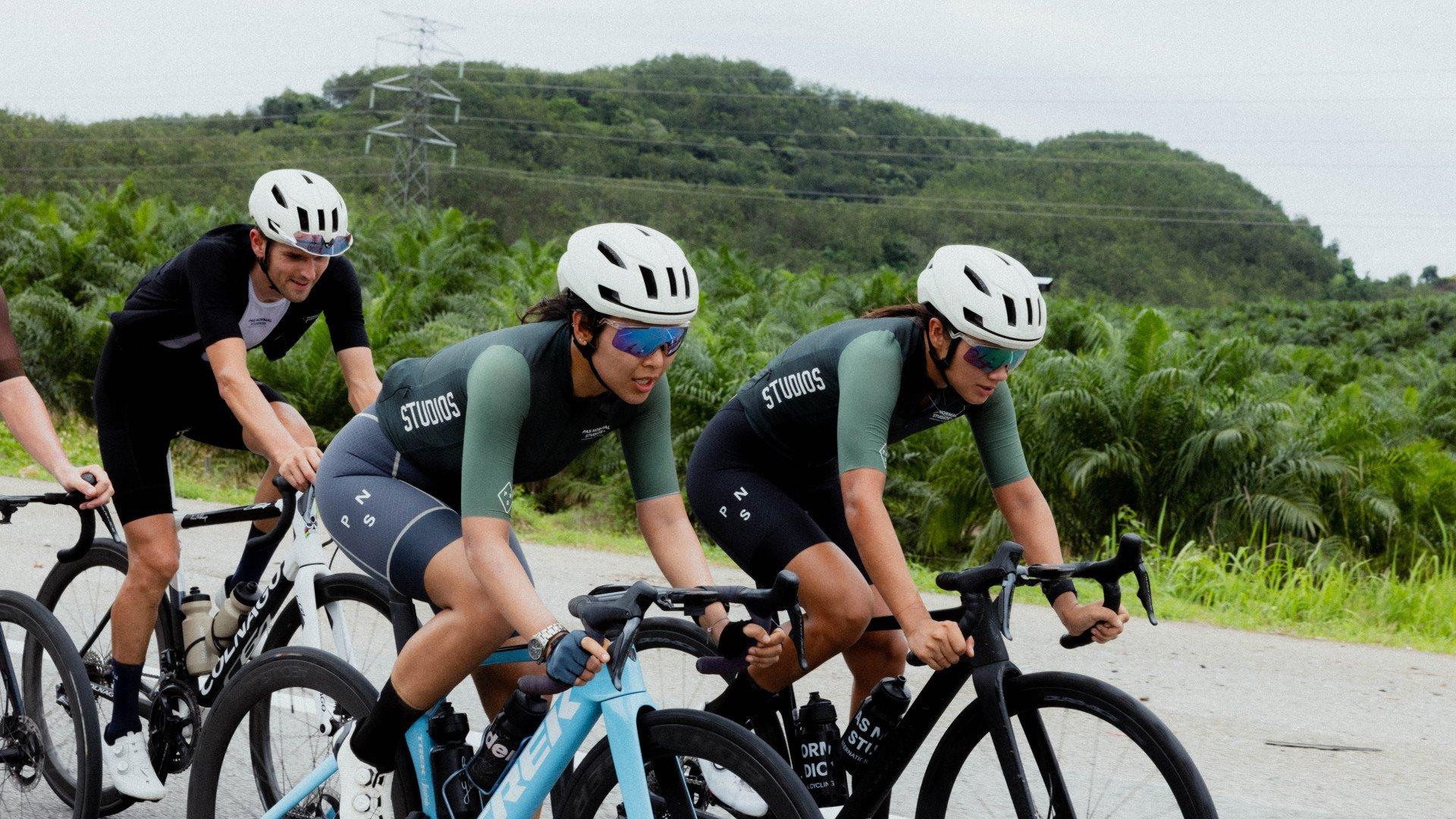 Three cyclists in aerodynamic position on time trial bikes wearing green and black skinsuit with white helmets on rural road