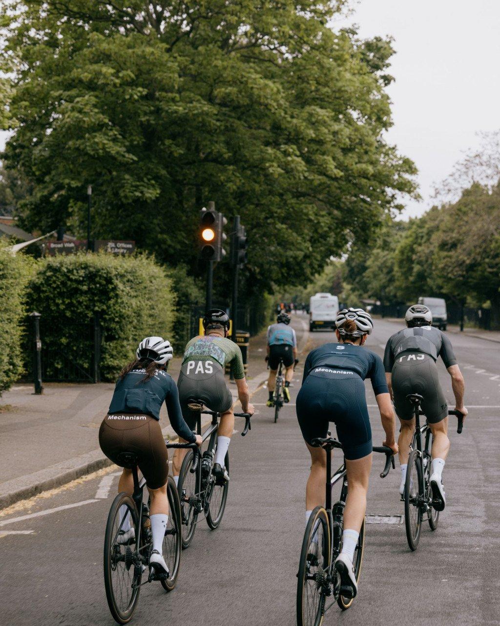 Group of cyclists in aerodynamic position wearing Mechanism collection jerseys and shorts on tree-lined road