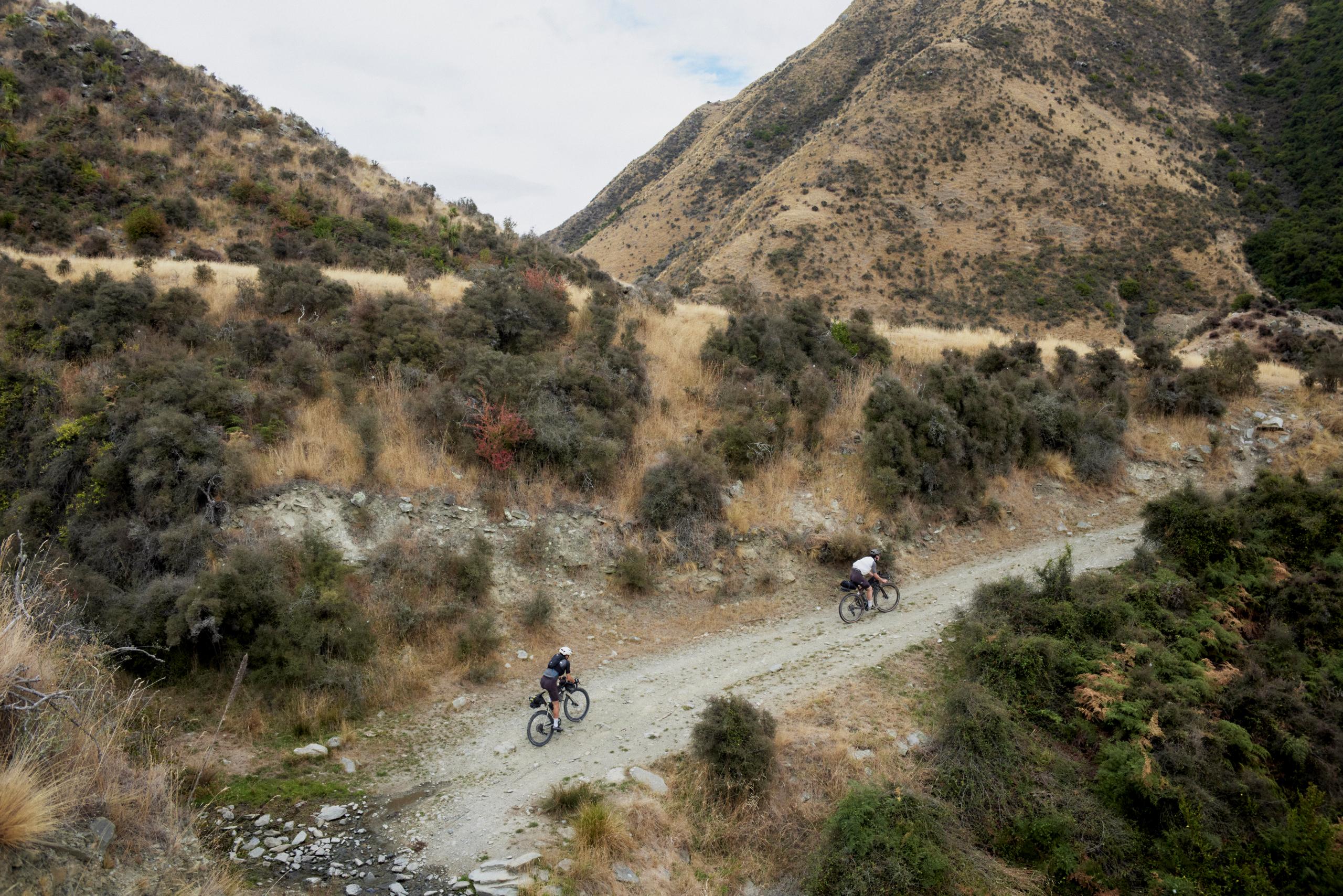 Two cyclists riding gravel bikes on dirt trail through mountainous terrain with golden hills and vegetation