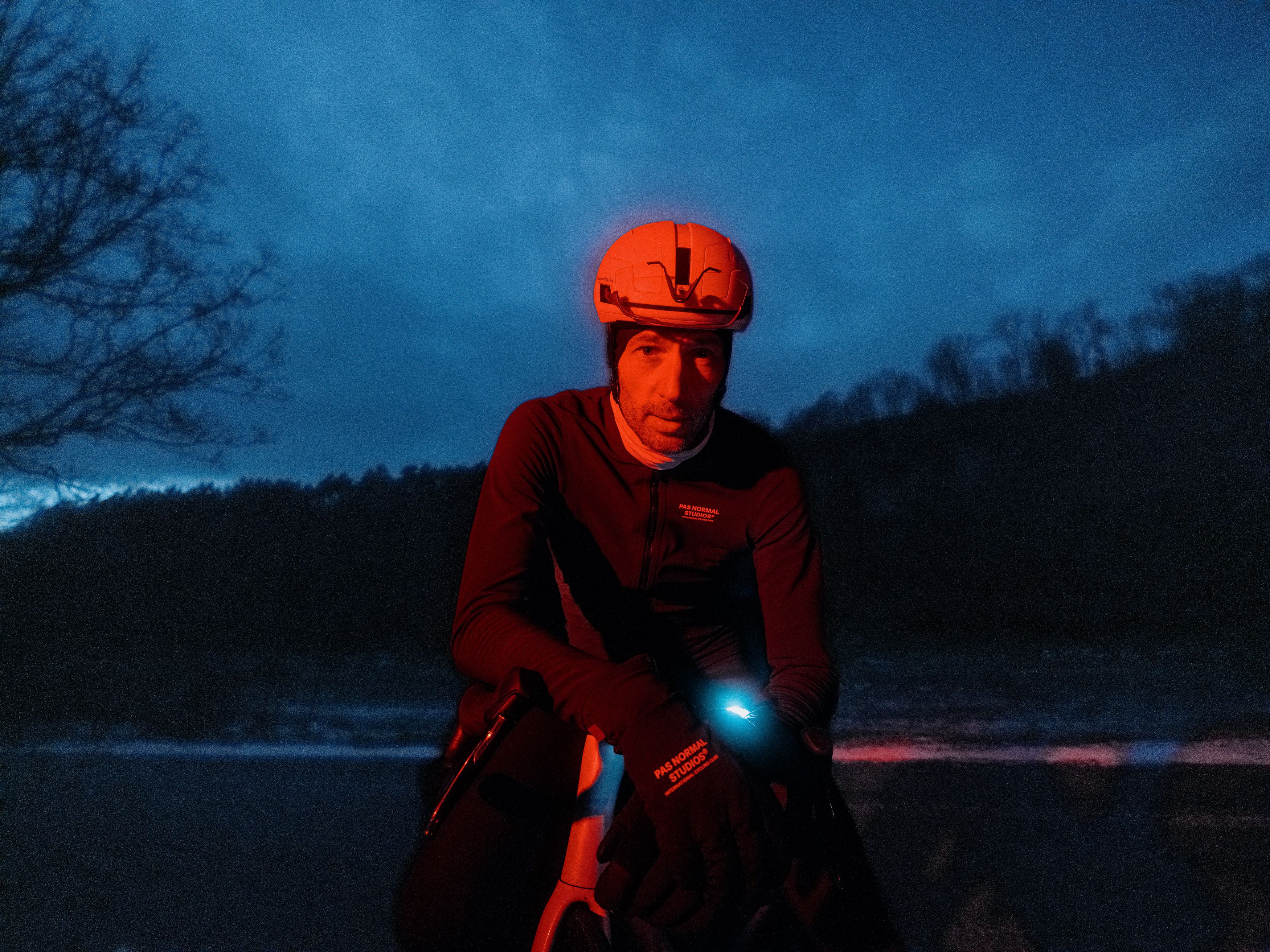 Model wearing orange cycling cap and dark jersey with chest branding cycling at dusk with front light