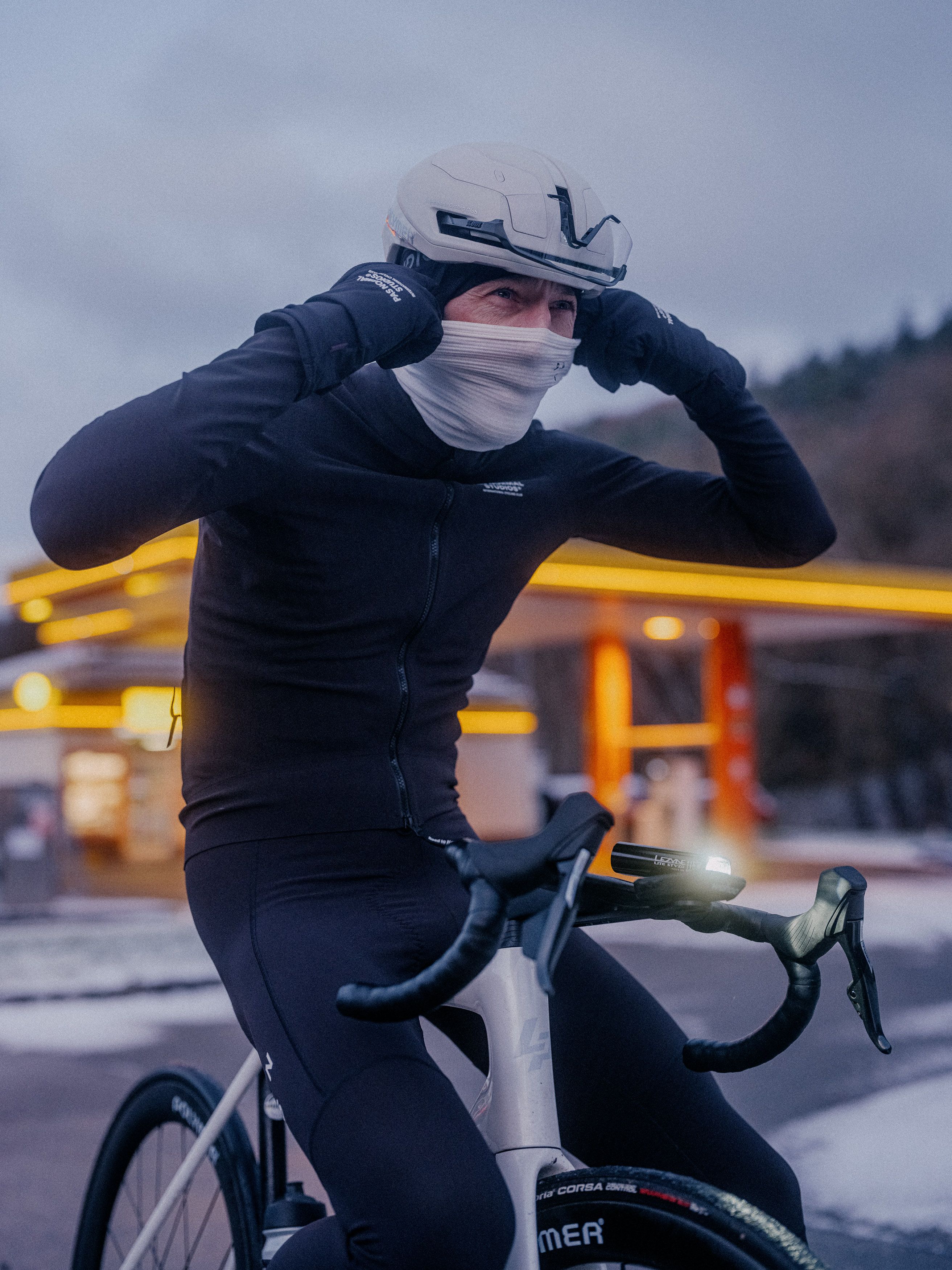 Model wearing black cycling skinsuit and white face covering on time trial bike in urban evening setting