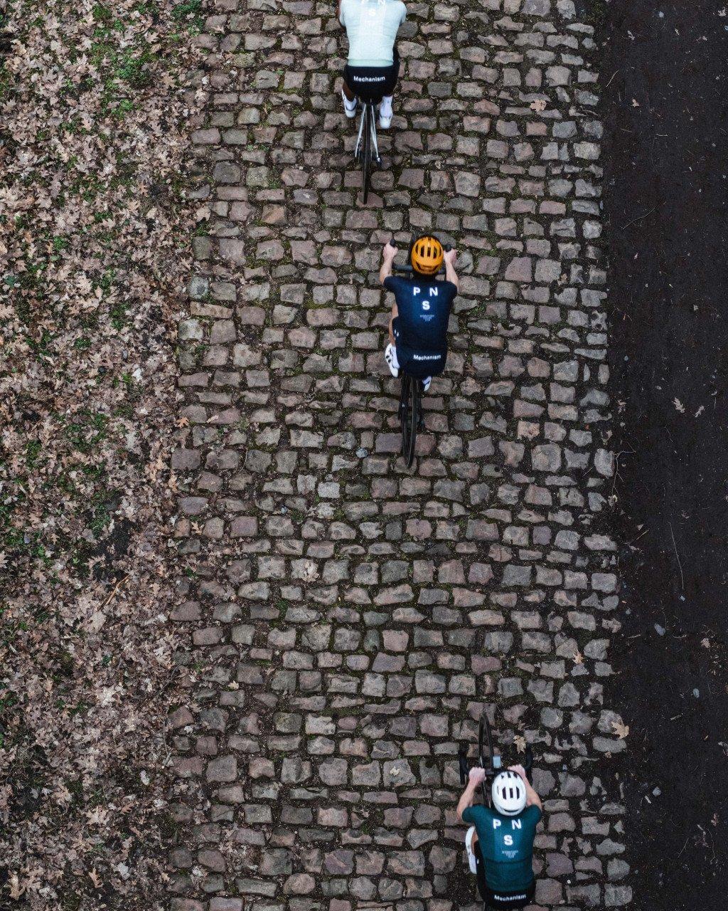 Three cyclists wearing Mechanism collection jerseys in navy and teal riding on cobblestone street, aerial view